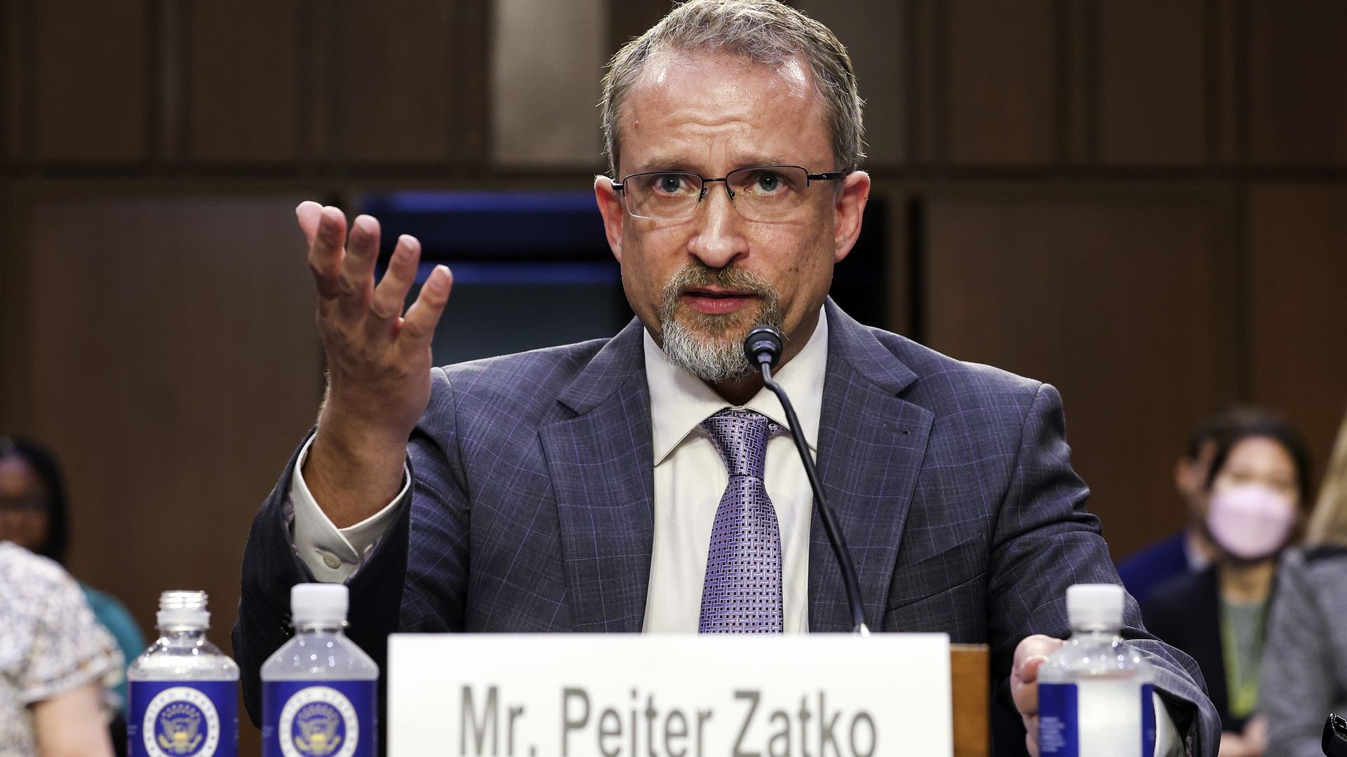 Photo of Peiter "Mudge" Zatko, arm gesturing, wearing a jacket and tie, with a name placard in front of him