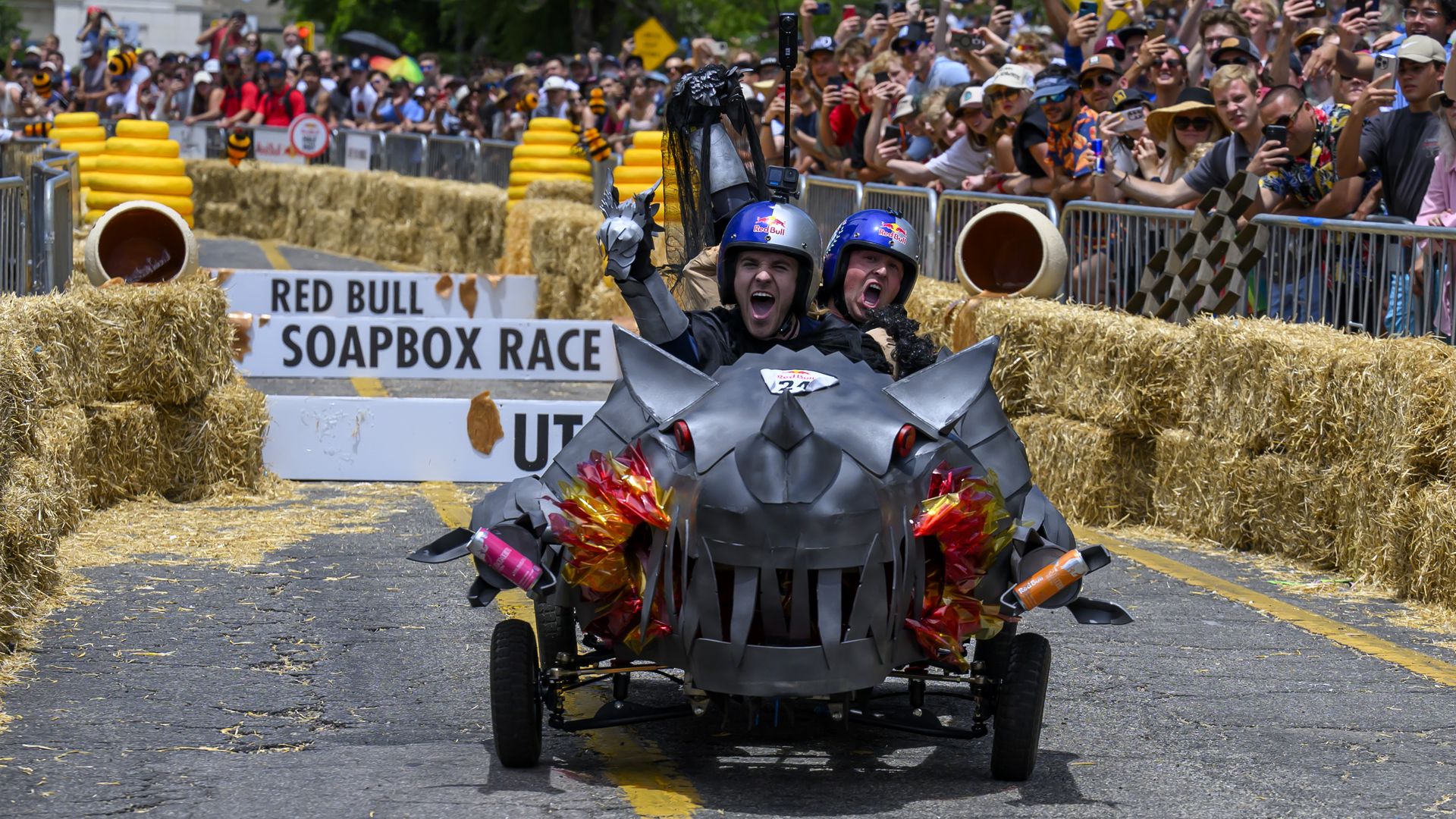 Two racers in a spiky black foam vehicle with helmets race down a street flanked by hay bales; a cheering crowd behind barriers holds up phones at the Red Bull Soapbox Race.