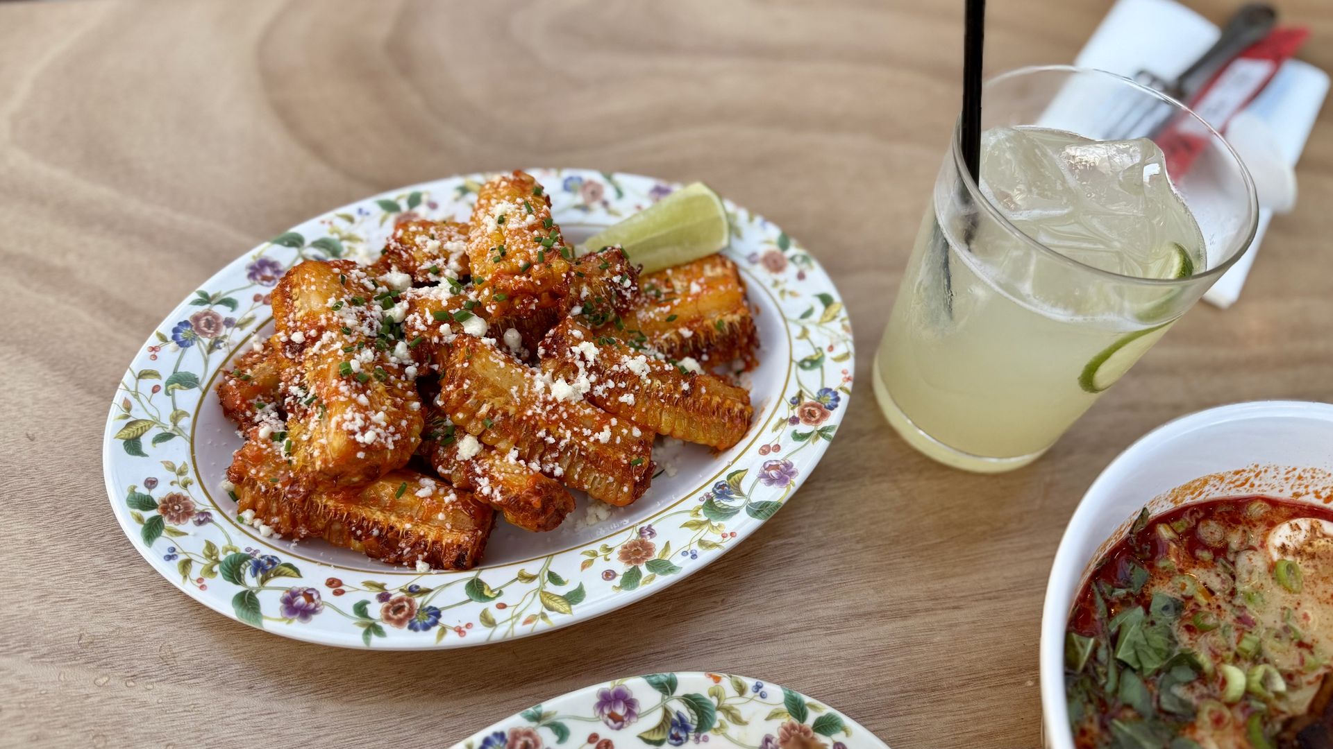 Plate of grilled corn covered with cheese and herbs, a lime wedge, a glass of iced lime drink with straw, and a bowl of spicy soup with herbs on a wooden table.