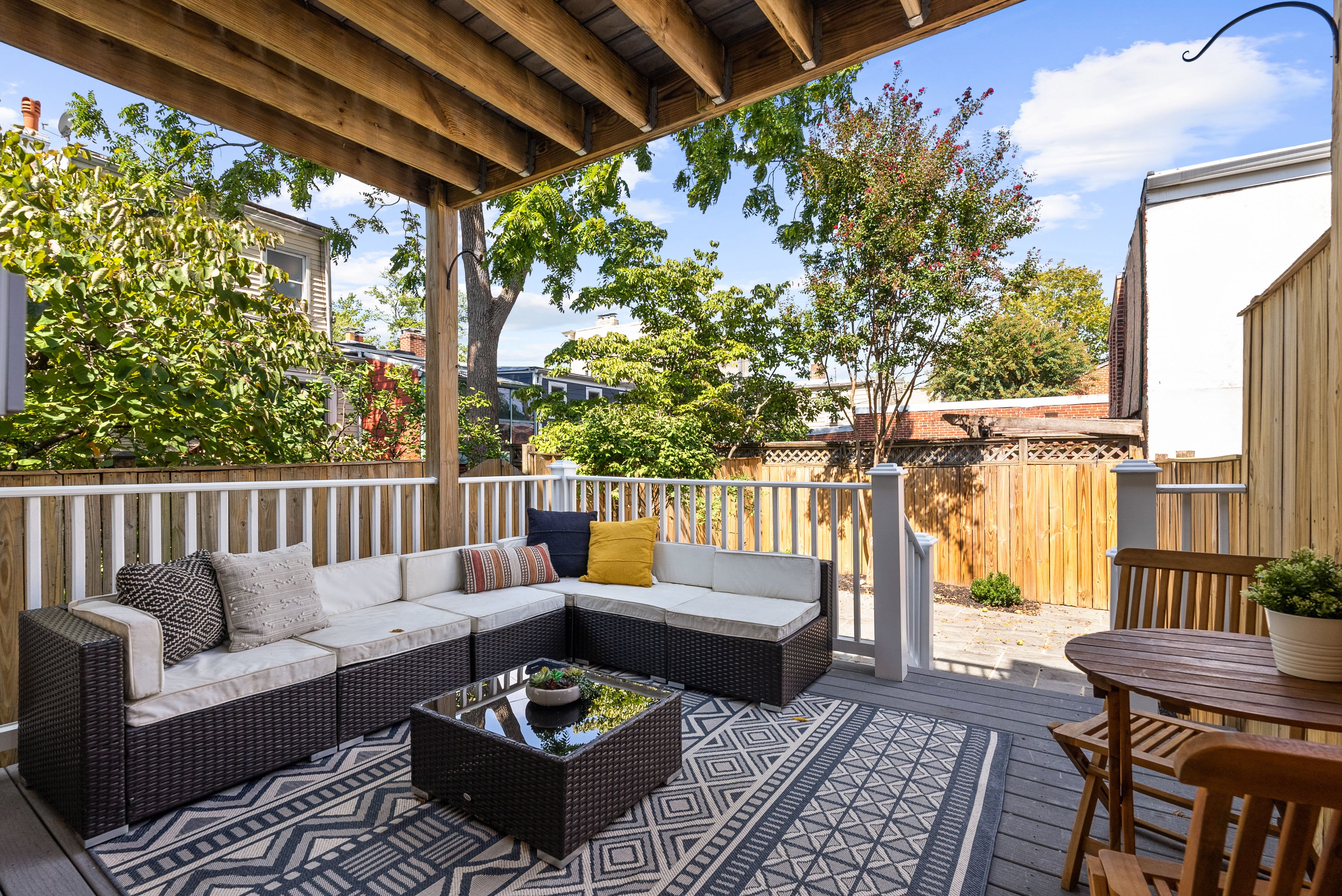 Covered patio with dark wicker sectional sofa with white cushions and patterned pillows, geometric rug, wooden table with chairs, surrounded by wooden fence and green trees under blue sky.