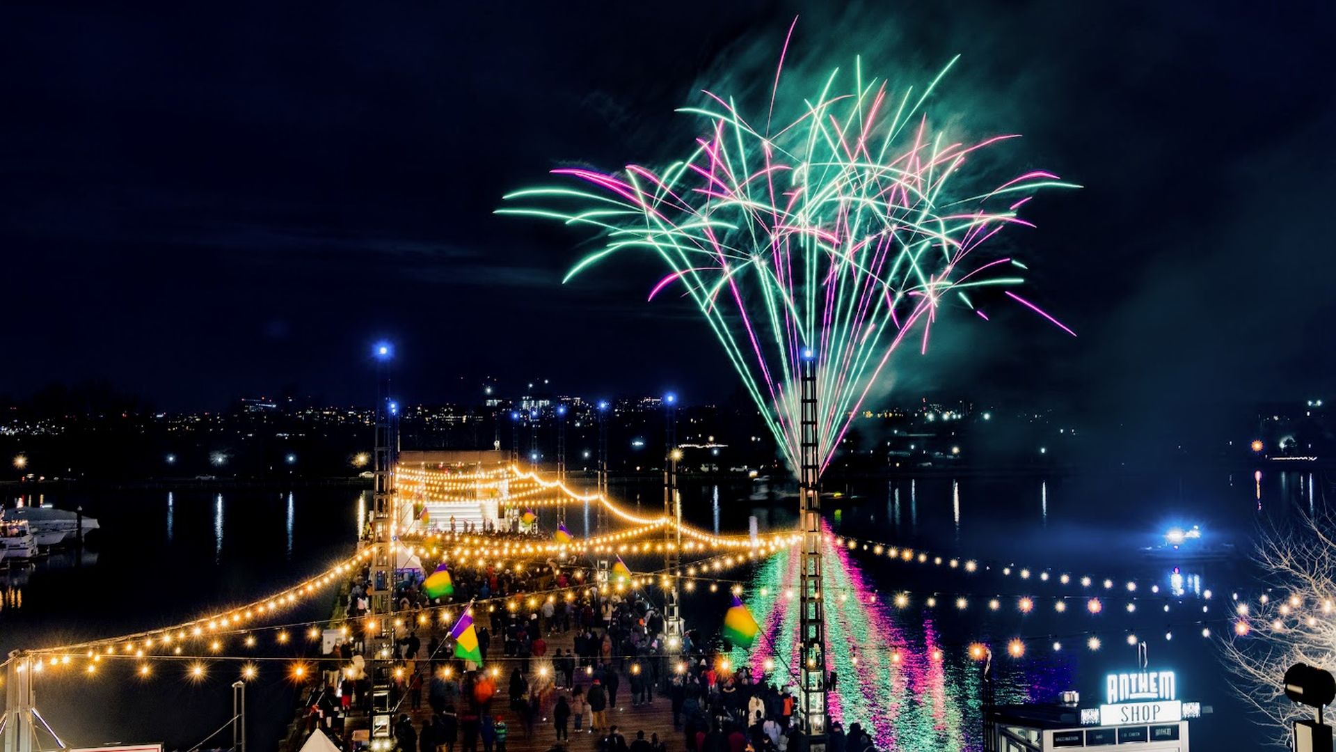 Nighttime fireworks in pink and green light up the sky and reflect on water, as a crowd gathers on a pier decorated with string lights and colorful flags near the Anthem Shop.