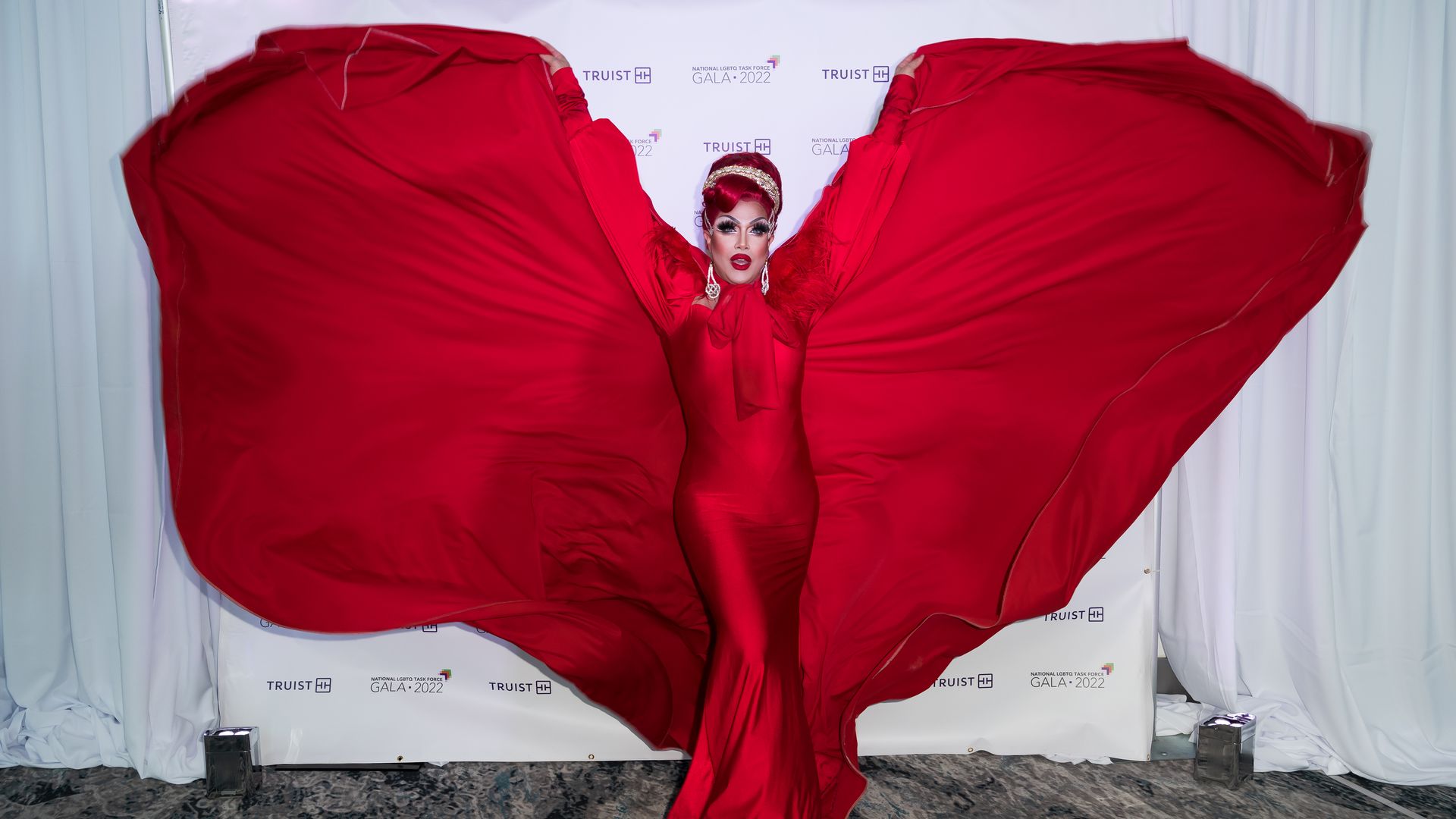 Person in a striking red gown with large flowing fabric wings extended, posing at the National LGBTQ Task Force Gala 2022 with a white backdrop. They wear bold makeup and earrings.