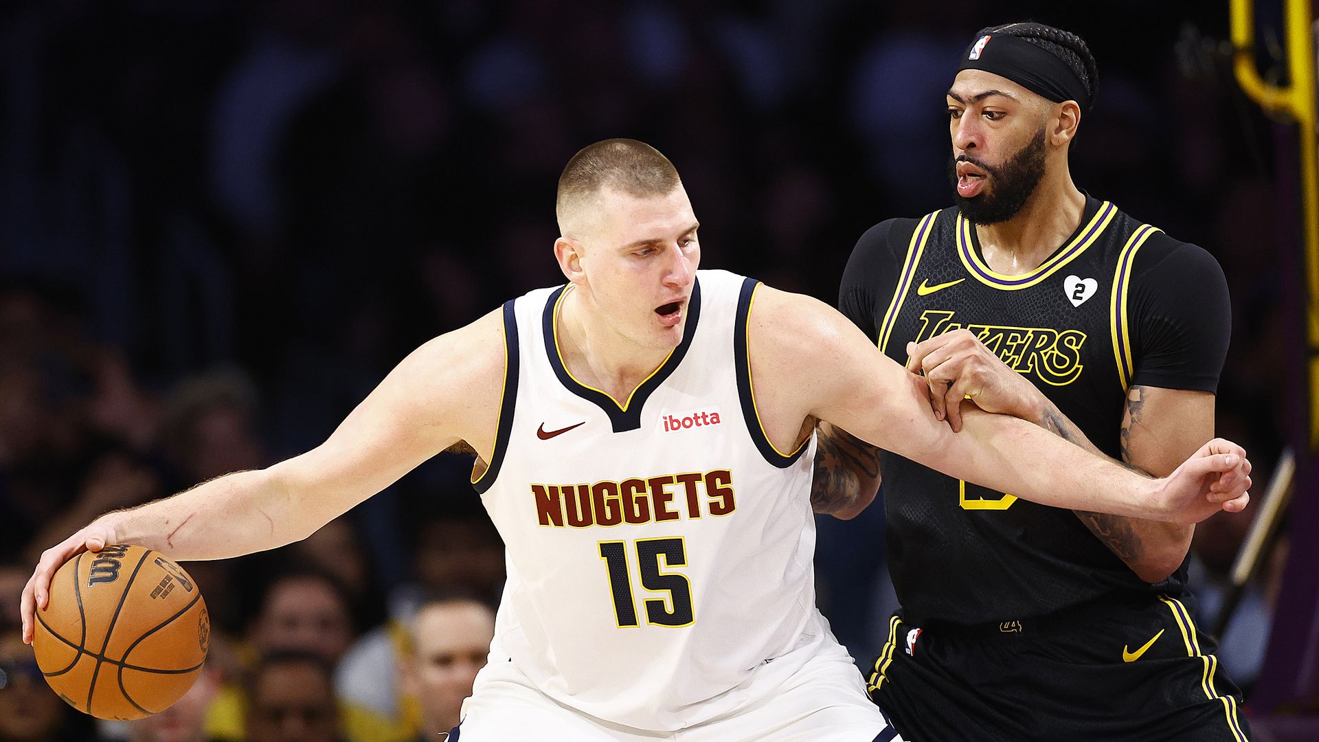 A man in a white jersey with the word NUGGETS and the number 15 on it holds an orange basketball while a man in a black jersey with yellow trimming tries to push up against the man.