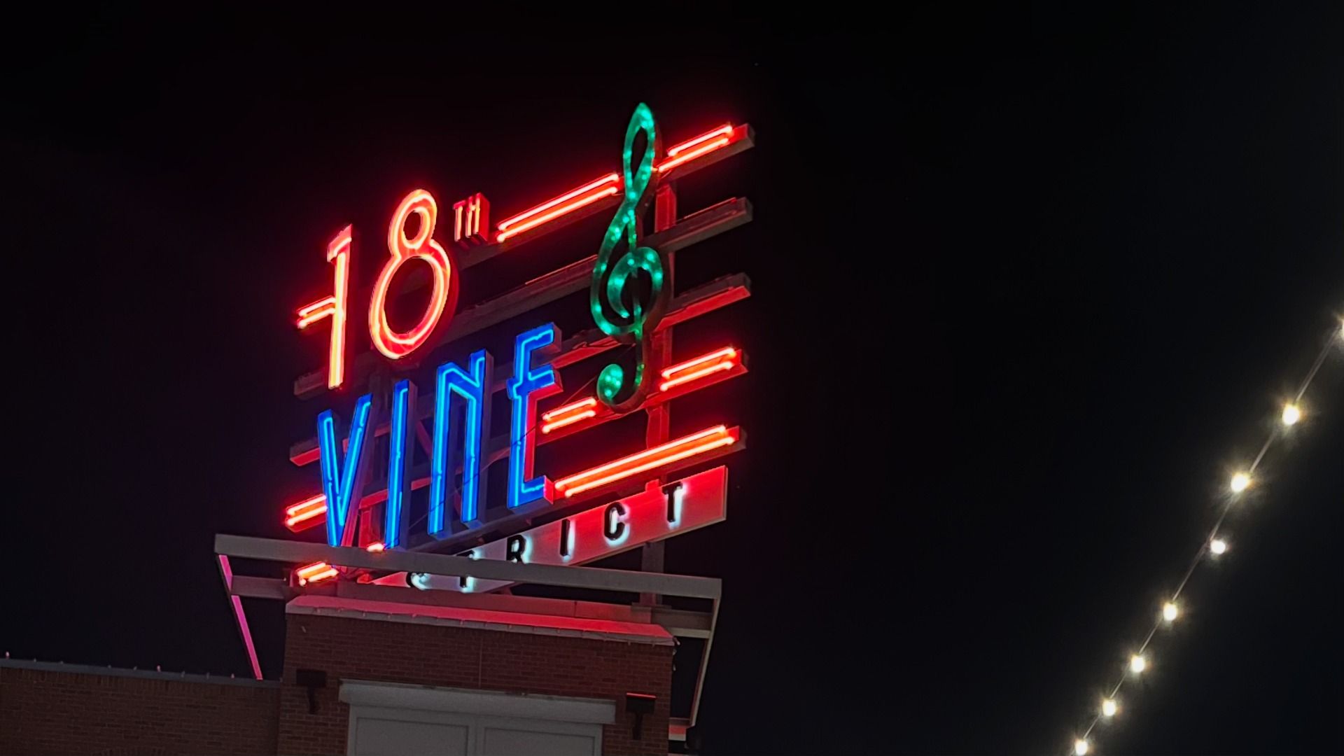 Nighttime neon sign reads "18th Vine" with a green treble clef over red and blue stripes; "DISTRICT" faint below, string lights to the right against a dark sky.