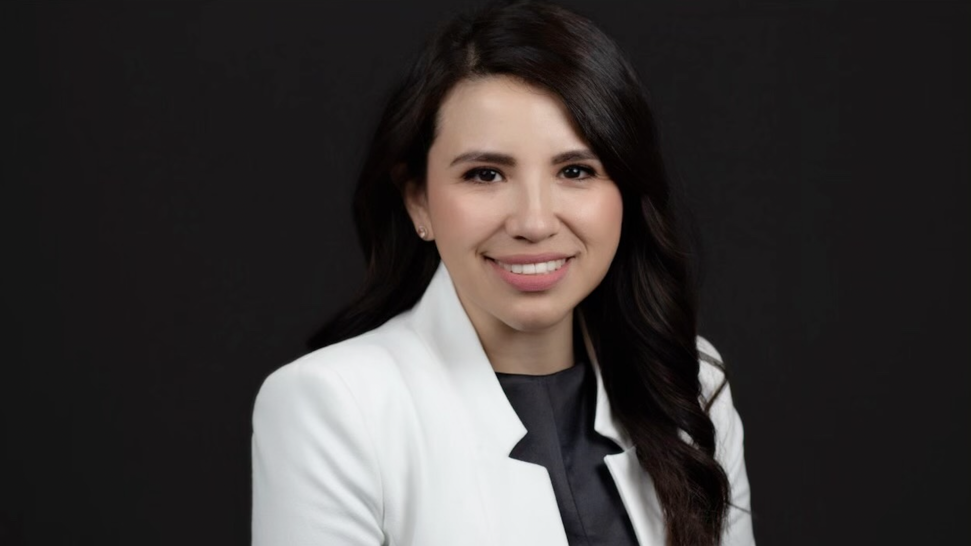 Cindy Nava poses for a photo with a black background. She is wearing a white blazer. 