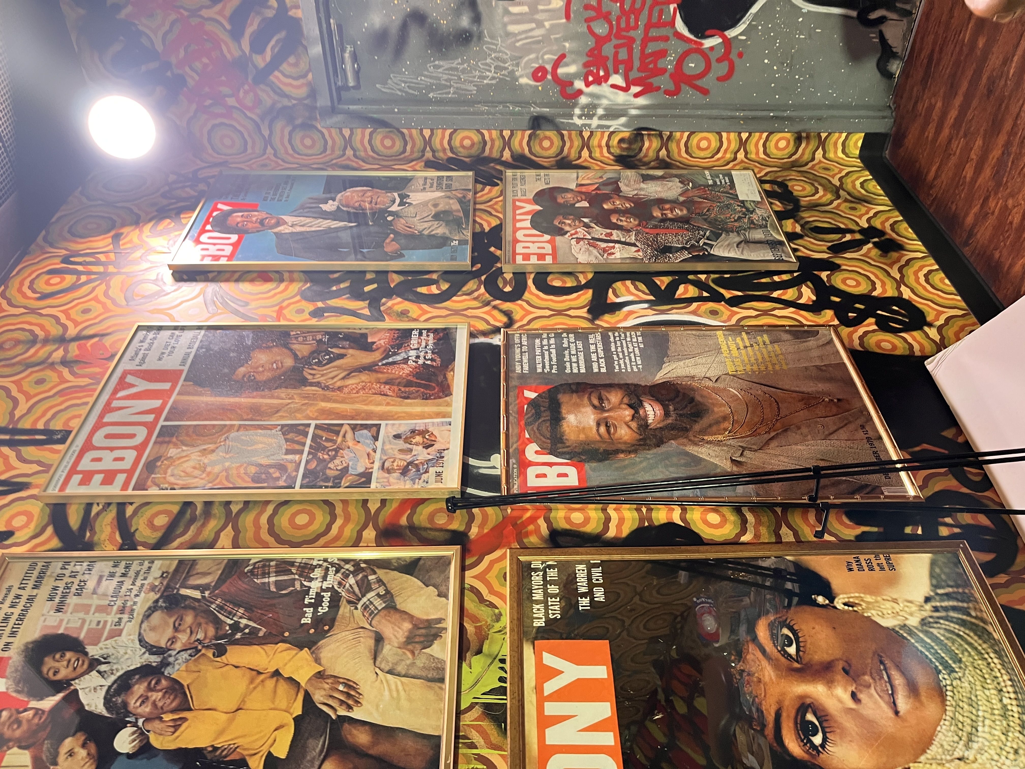 Retro room with psychedelic yellow and orange wallpaper. Framed magazine covers line the wall; a gray graffiti-covered cabinet sits center as a bright round lamp on the left lights a wooden floor.