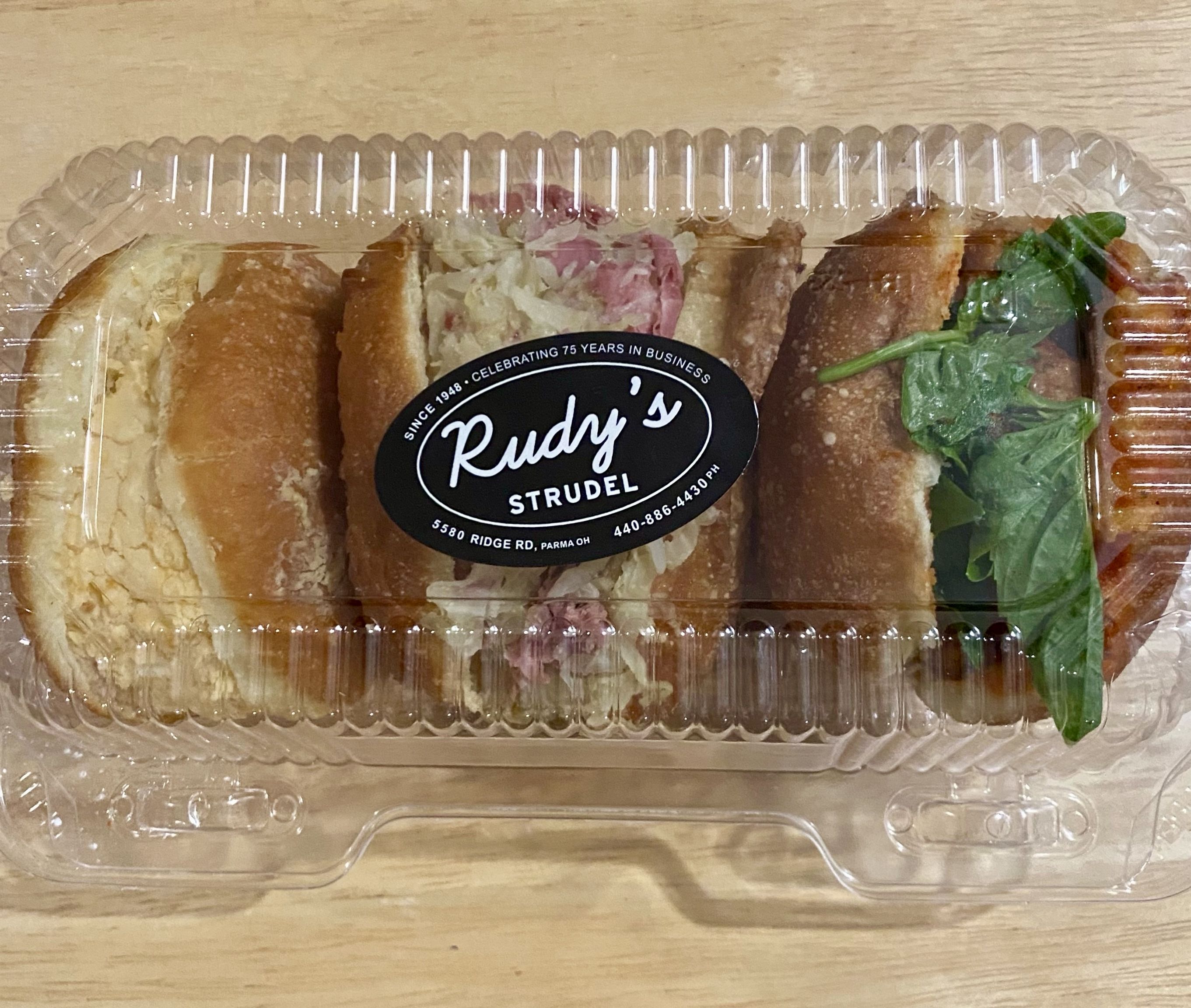Plastic container with five different strudels from Rudy's Strudel, including an egg salad, a pink meat with sauerkraut, a spinach, and others, on a wooden surface.