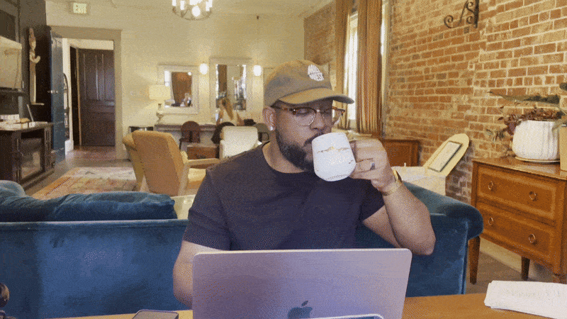 Man wearing a cap and glasses drinks from a white mug while working on a laptop in a cozy room with exposed brick walls, blue couch, wooden furniture, and warm lighting.