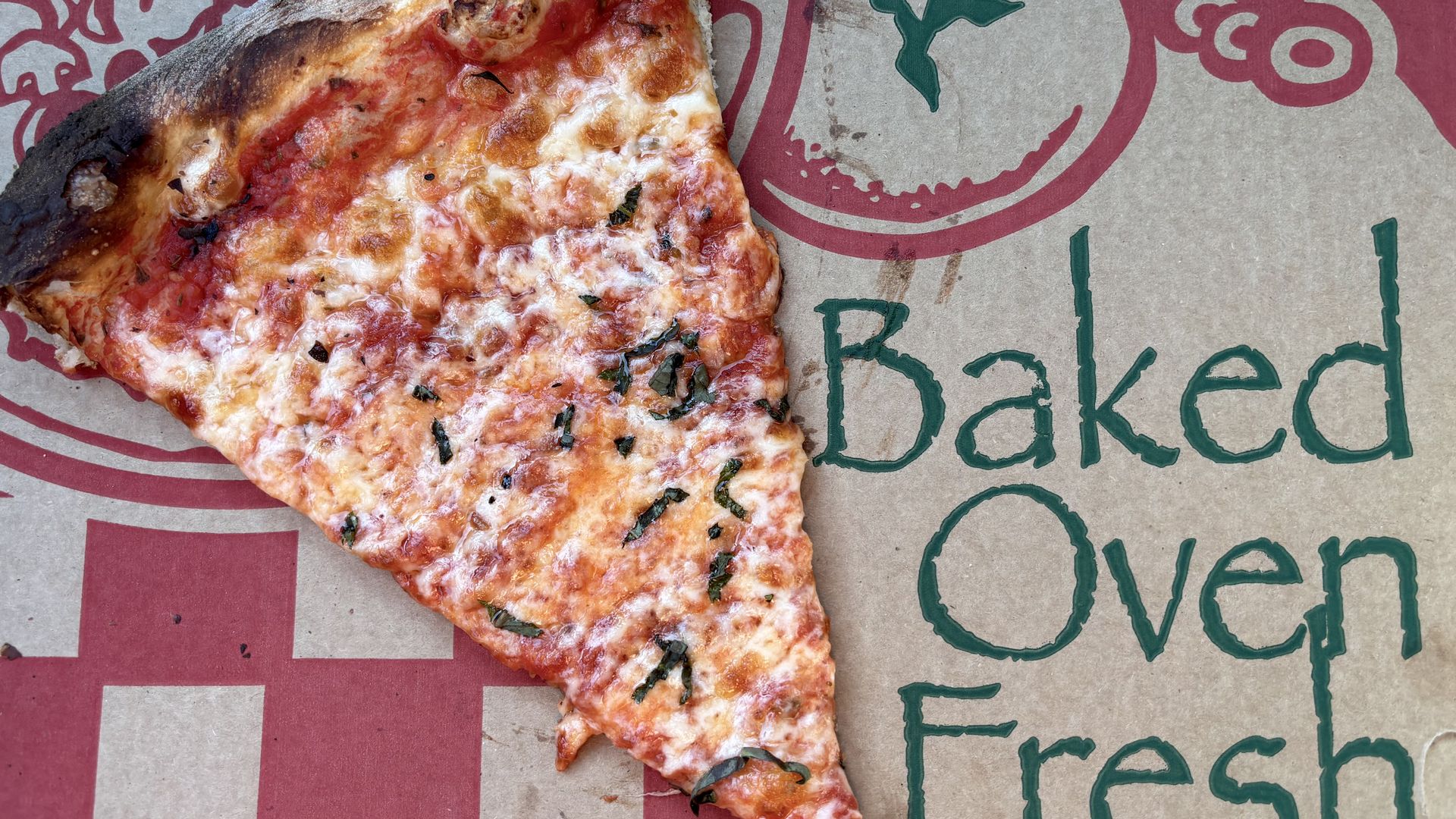 Close-up of a cheese pizza slice with melted cheese and herbs on a cardboard pizza box with red and green printing; the crust on the left is dark and charred.