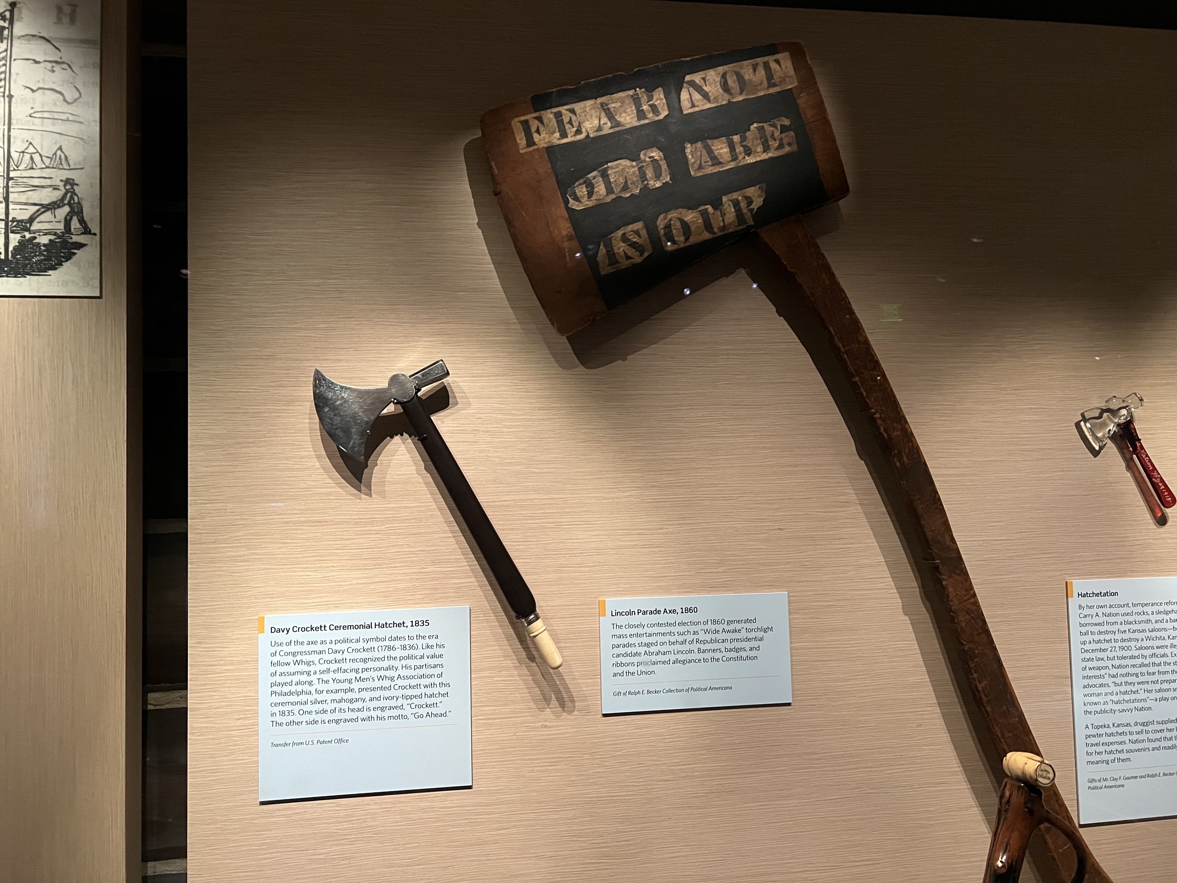 Display of three vintage axes in a museum with information plaques, including a large wooden Lincoln Parade Axe from 1860 with black text reading "FEAR NOT OLD AGE IS OUR".