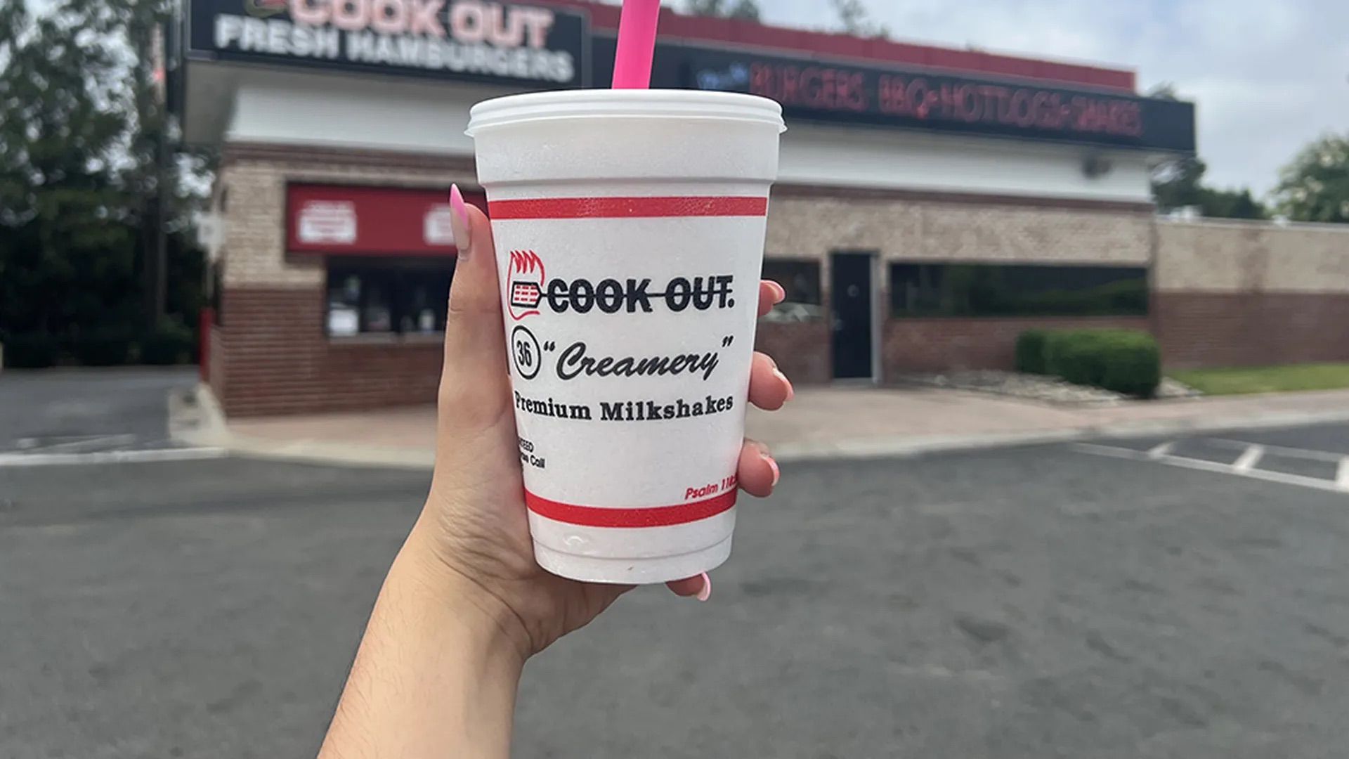 Hand holding a white milkshake cup with red stripes and pink straw in front of a Cook Out fast food restaurant with brick exterior and signage advertising burgers, BBQ, hotdogs, and shakes.