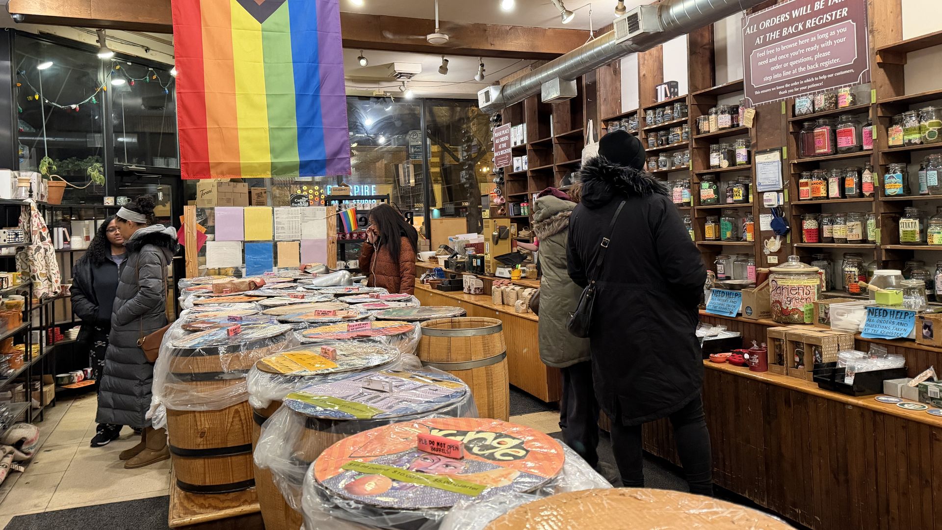 Interior of a store with wooden barrels covered in plastic and colorful labels, people shopping, shelves filled with jars, and a large Progress Pride flag hanging from the ceiling.