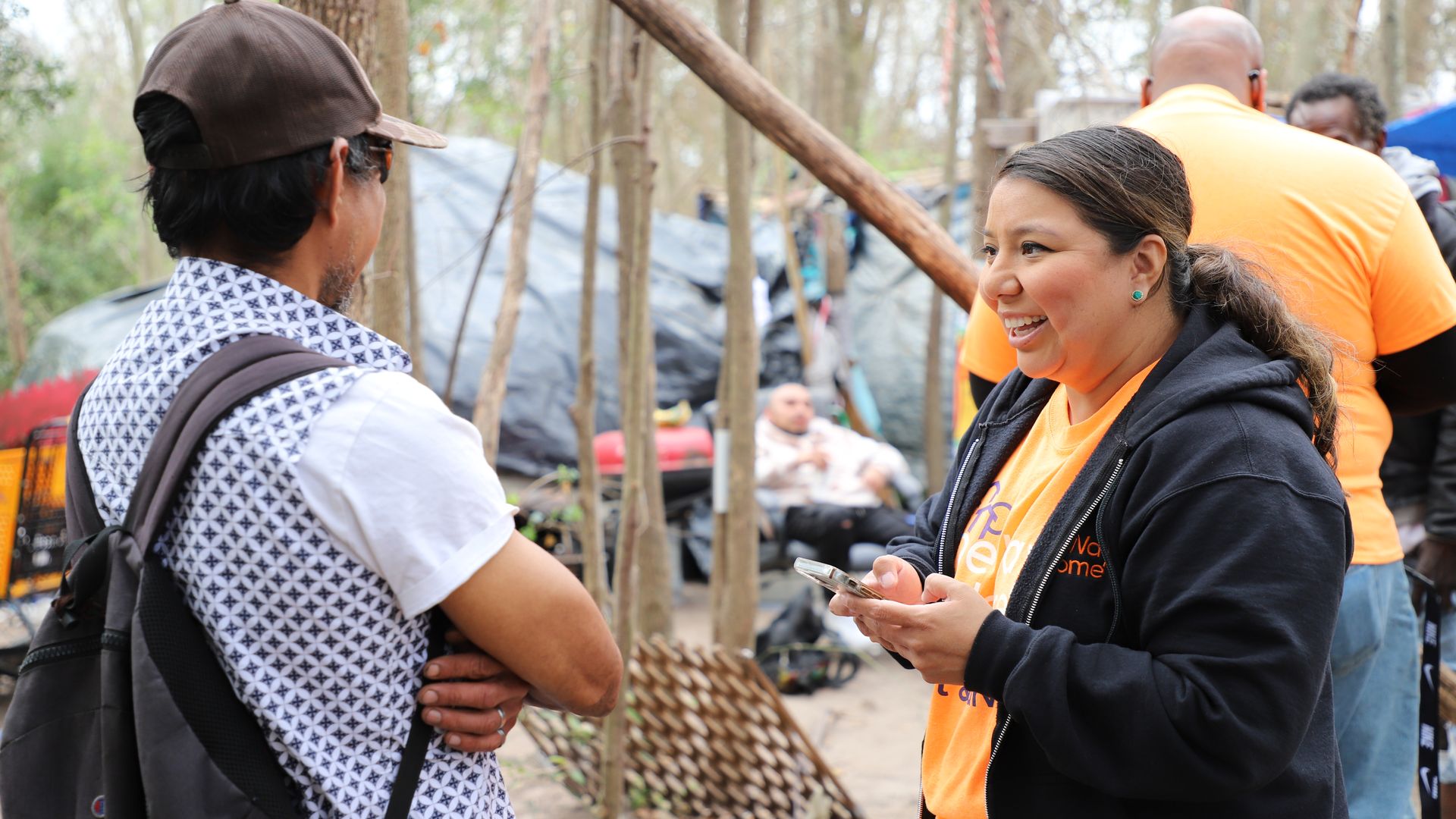 Two people chat outdoors in a wooded area; a man wearing a brown cap and patterned shirt faces a smiling woman in an orange shirt and black jacket. Background shows trees and volunteers.