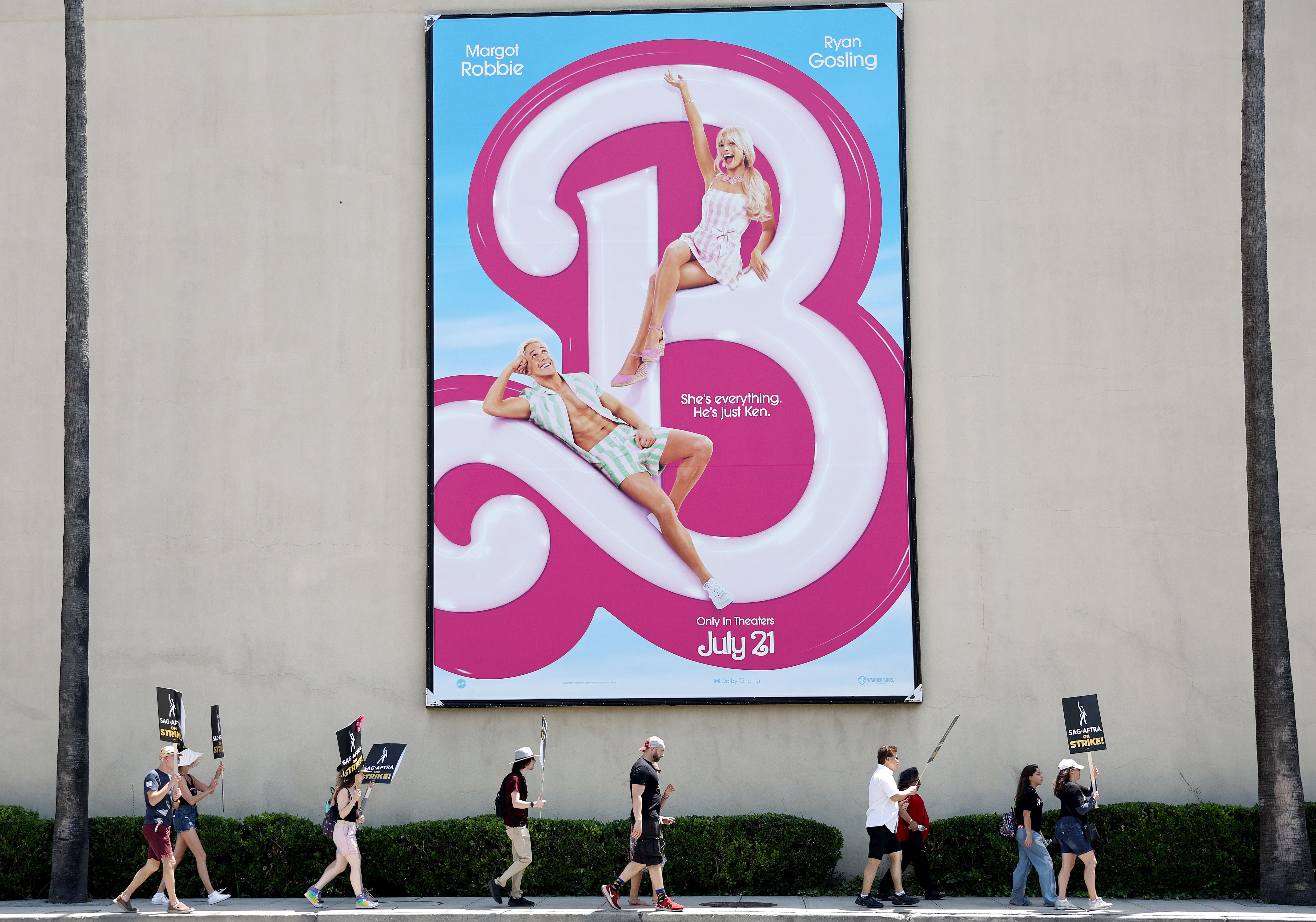 An image of a giant Barbie poster with SAG protestors marching in the foreground.