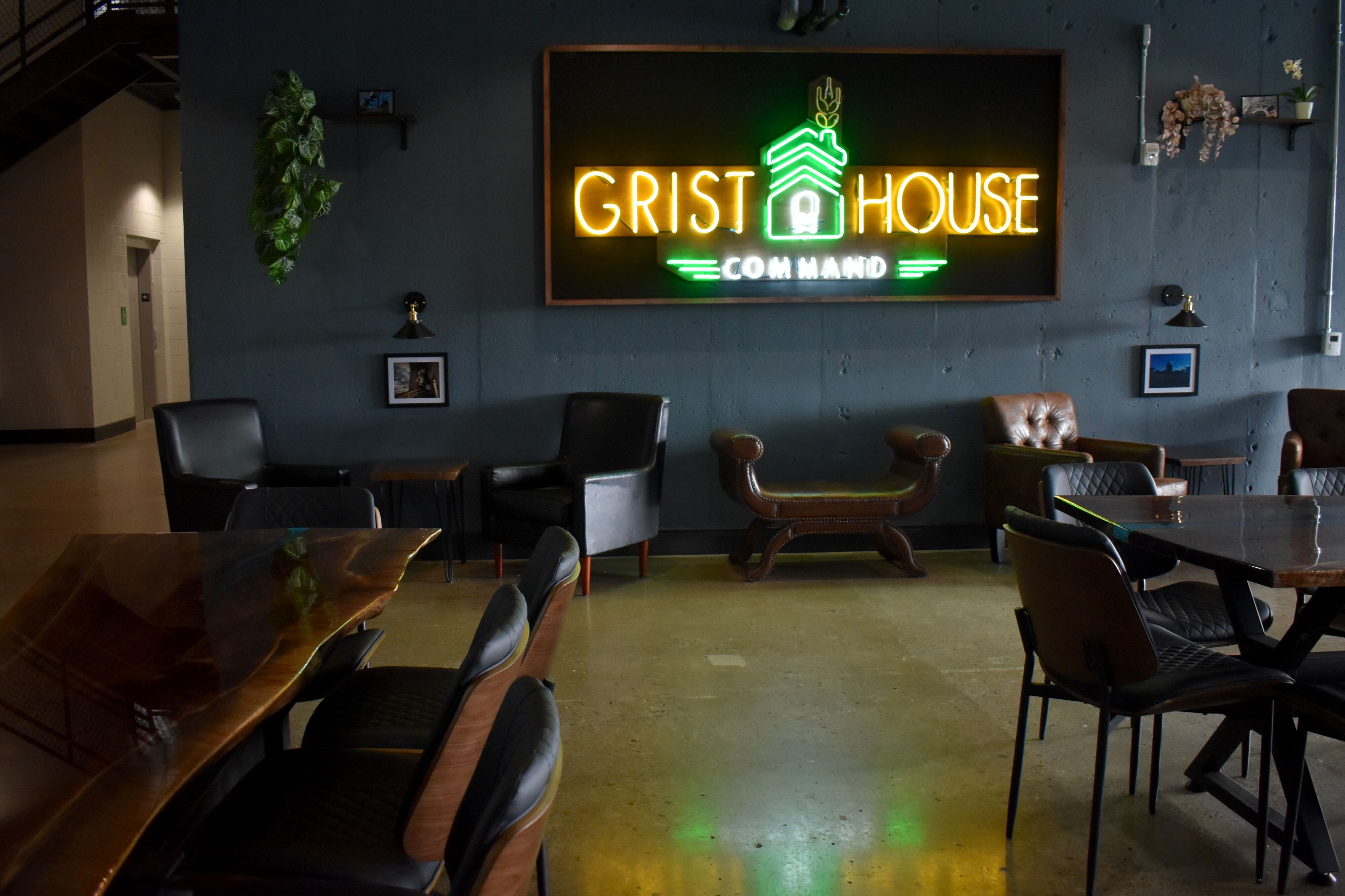 Interior of a lounge with dark blue walls, leather chairs, wooden tables, and a neon sign reading GRIST HOUSE COMMAND in yellow and green.