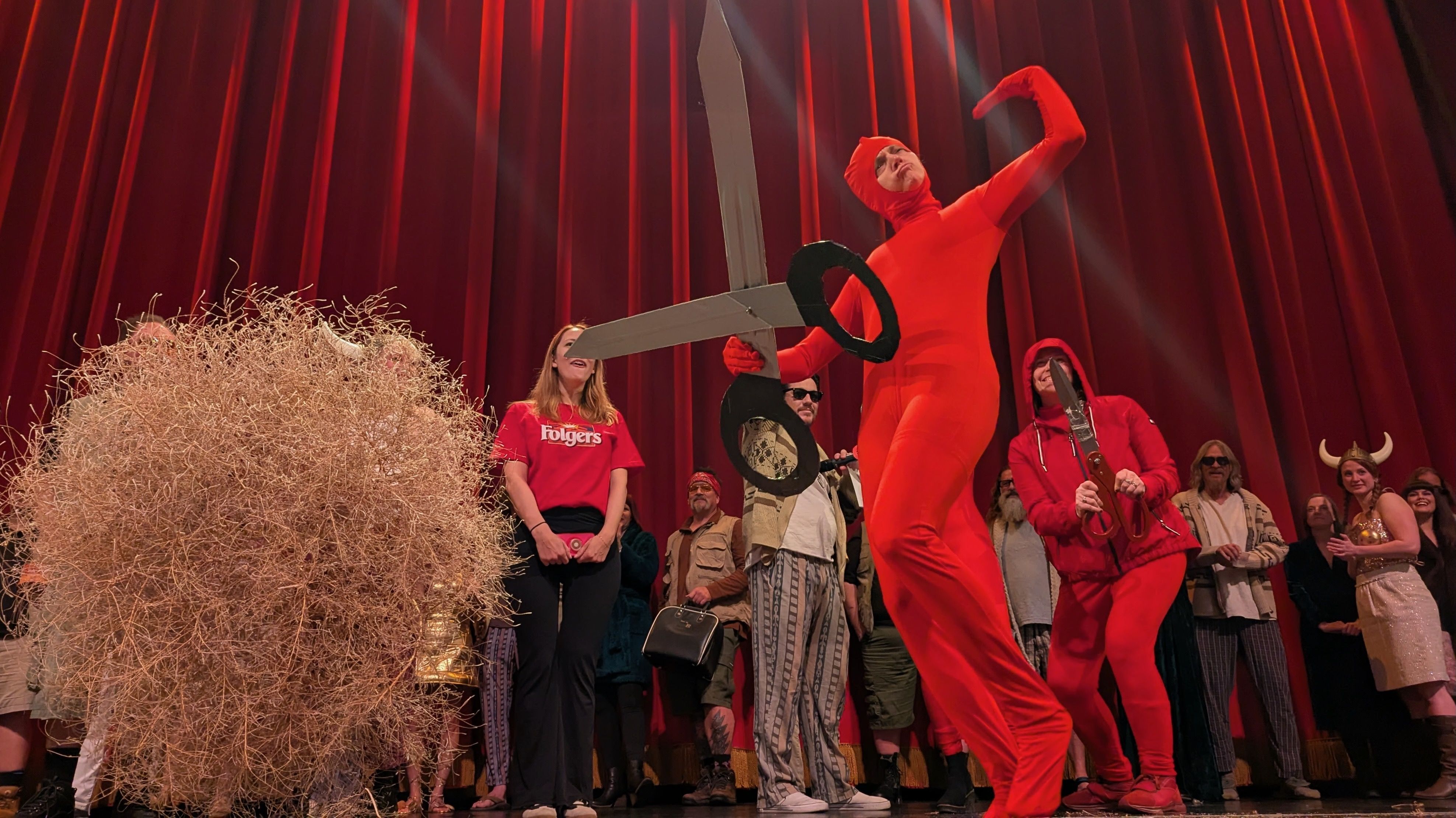 A woman in a red jumpsuit carrying giant scissors on a stage near a sagebrush and others in costumes.