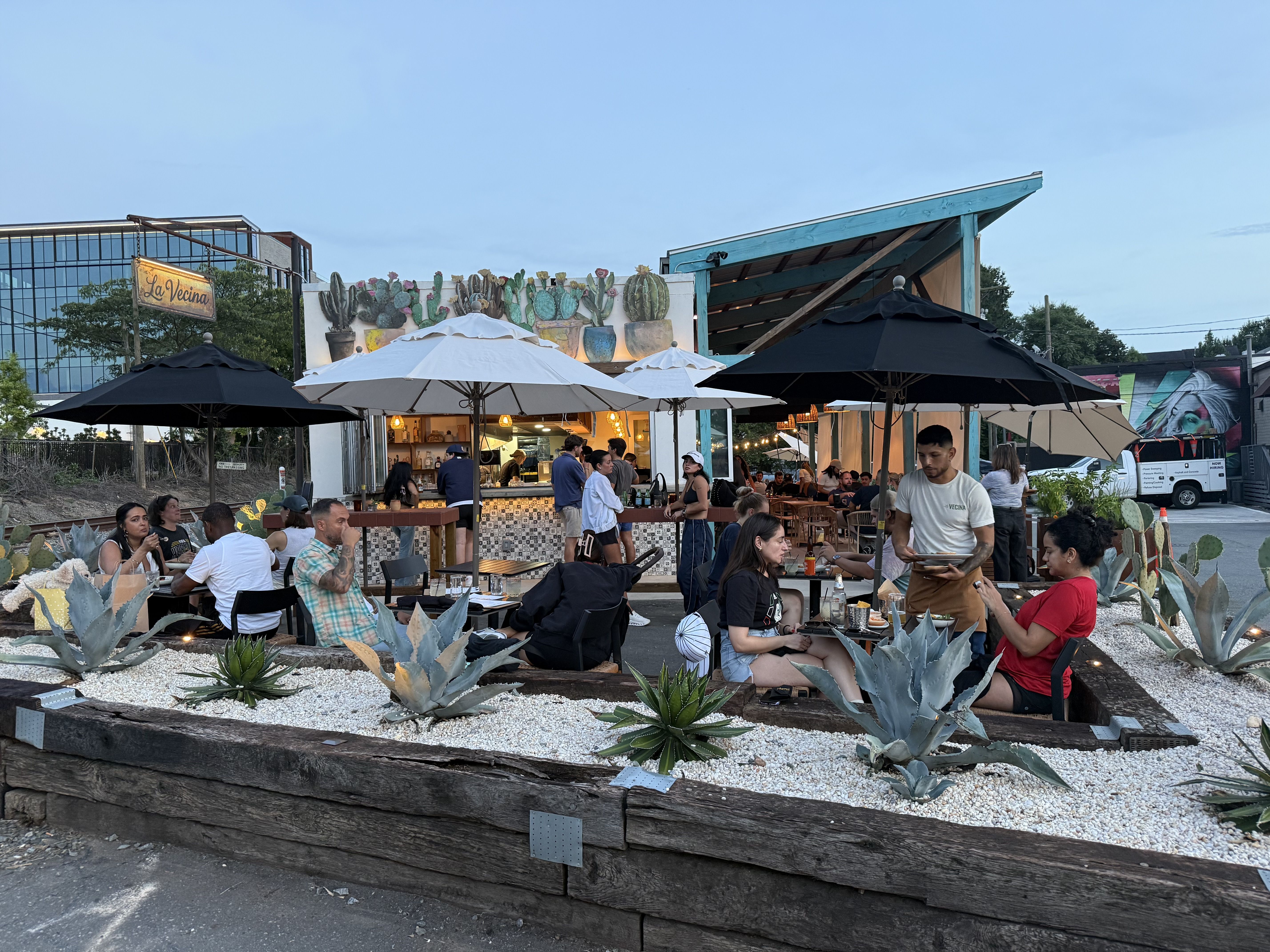 Outdoor dining scene with many people beneath white and black umbrellas, a La Vecina sign on the left, a colorful cactus mural, foreground succulents, and a teal-roofed shelter under a clear blue sky.