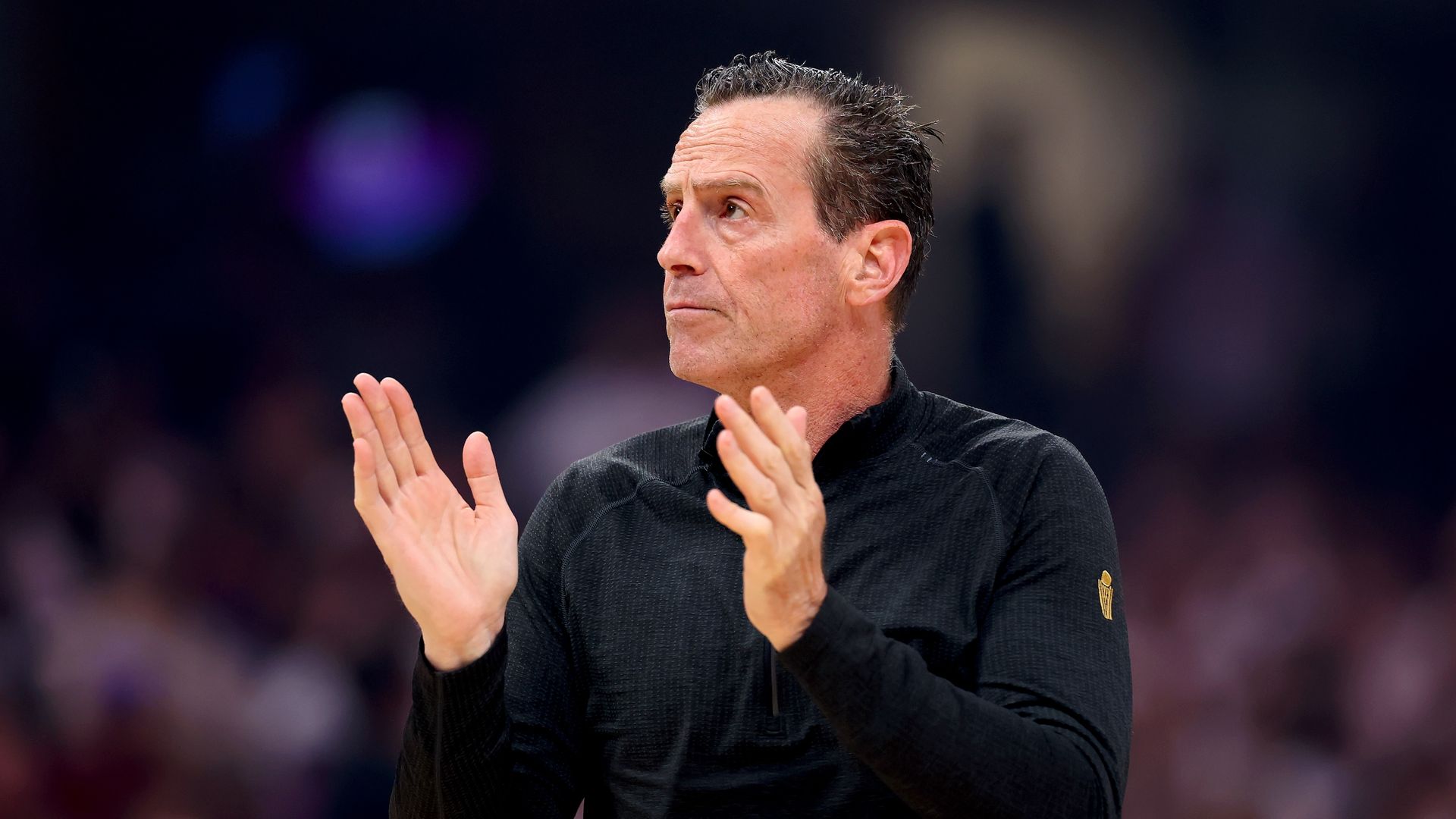 Side view of a middle-aged man in a black athletic shirt with a small gold crest on the sleeve, clapping his hands and looking to the left in a dim sports arena.
