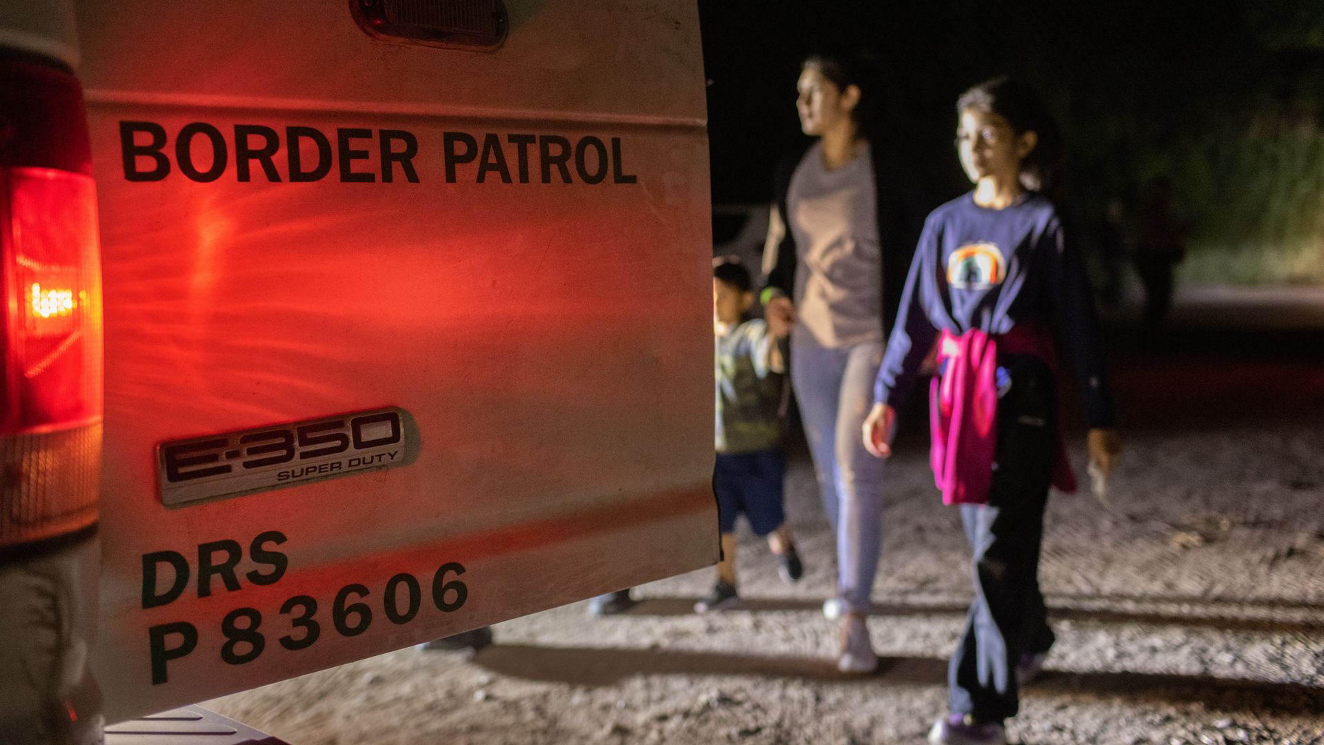 Photo of an open vehicle door that says "Border Patrol" and a group of migrants walking toward it