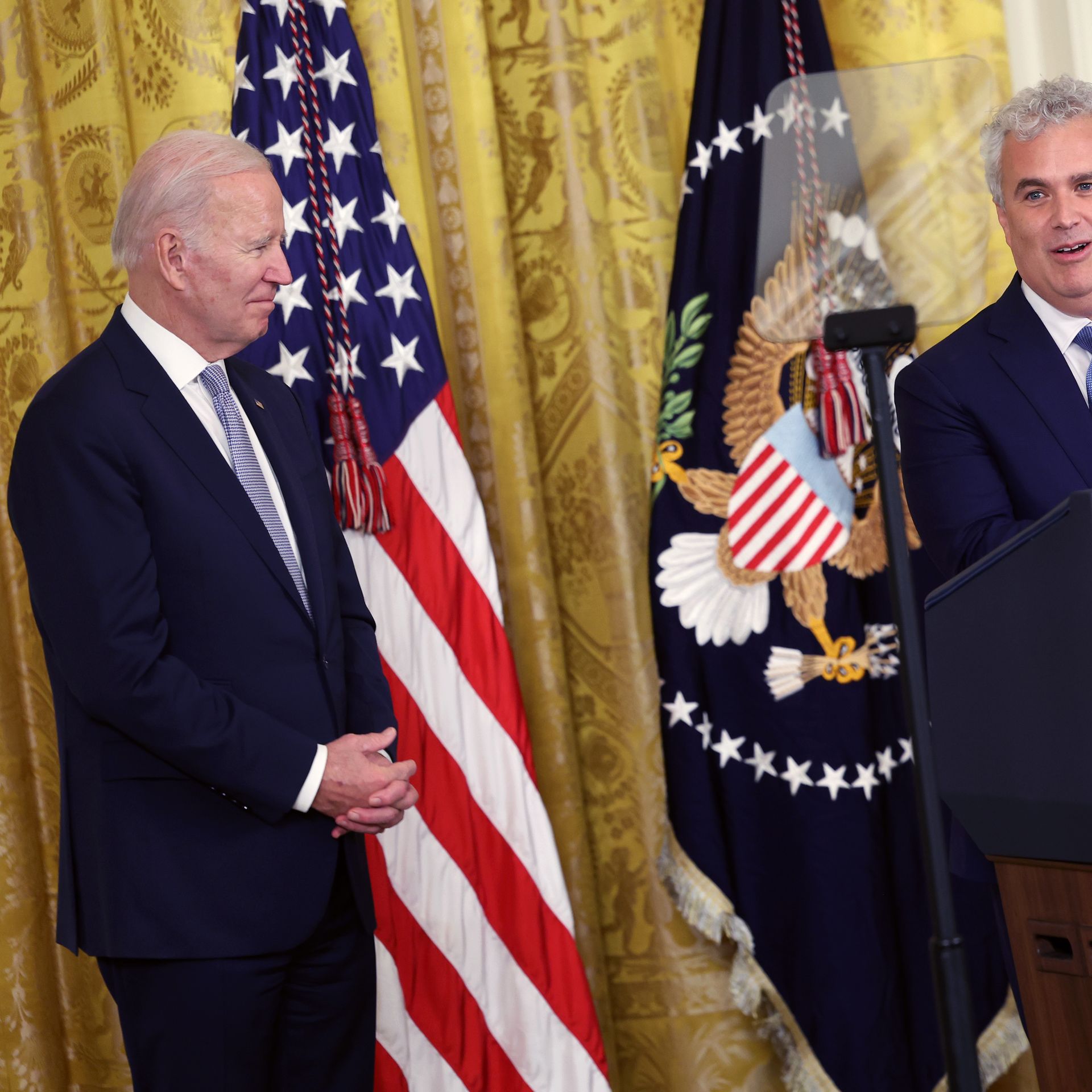 Joe Biden looks onto Jeff Zients before flags and the golden drapes of the east room of the White House