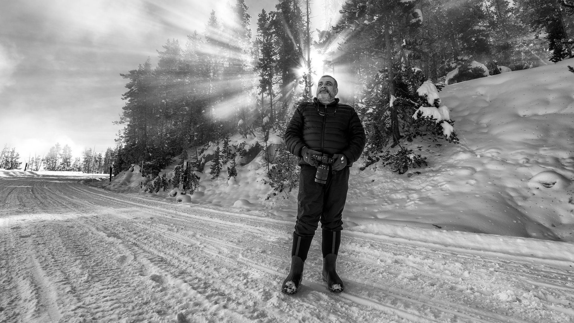 Black-and-white photo of a man in a snowy forest, wearing a puffer jacket and holding a camera, with sun rays streaming through tall evergreen trees behind him.