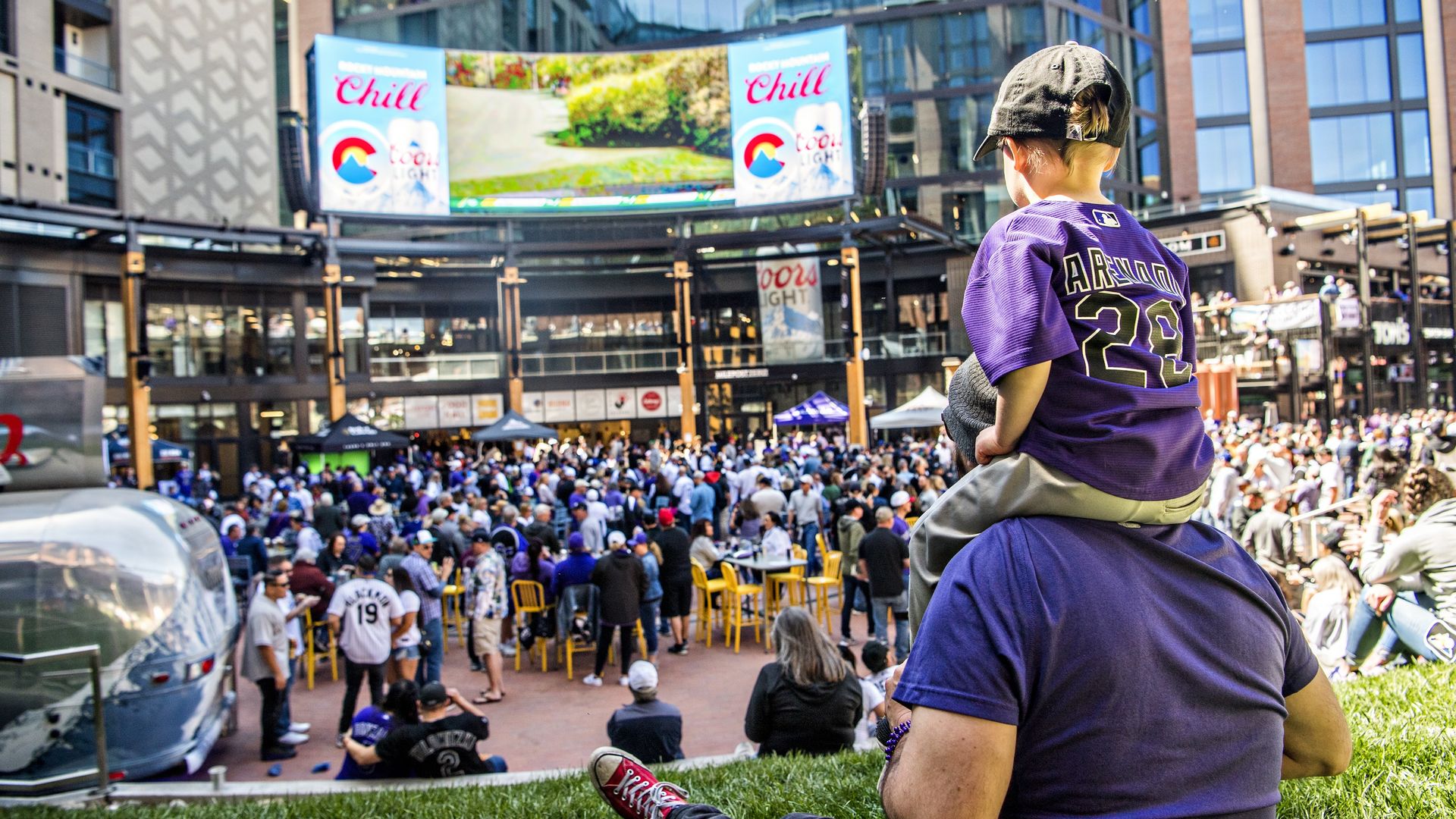 Adult in a purple jersey with the number 23 carries a child on their shoulders in the foreground, overlooking a crowded urban plaza with a large screen, banners, and yellow chairs among modern buildings.