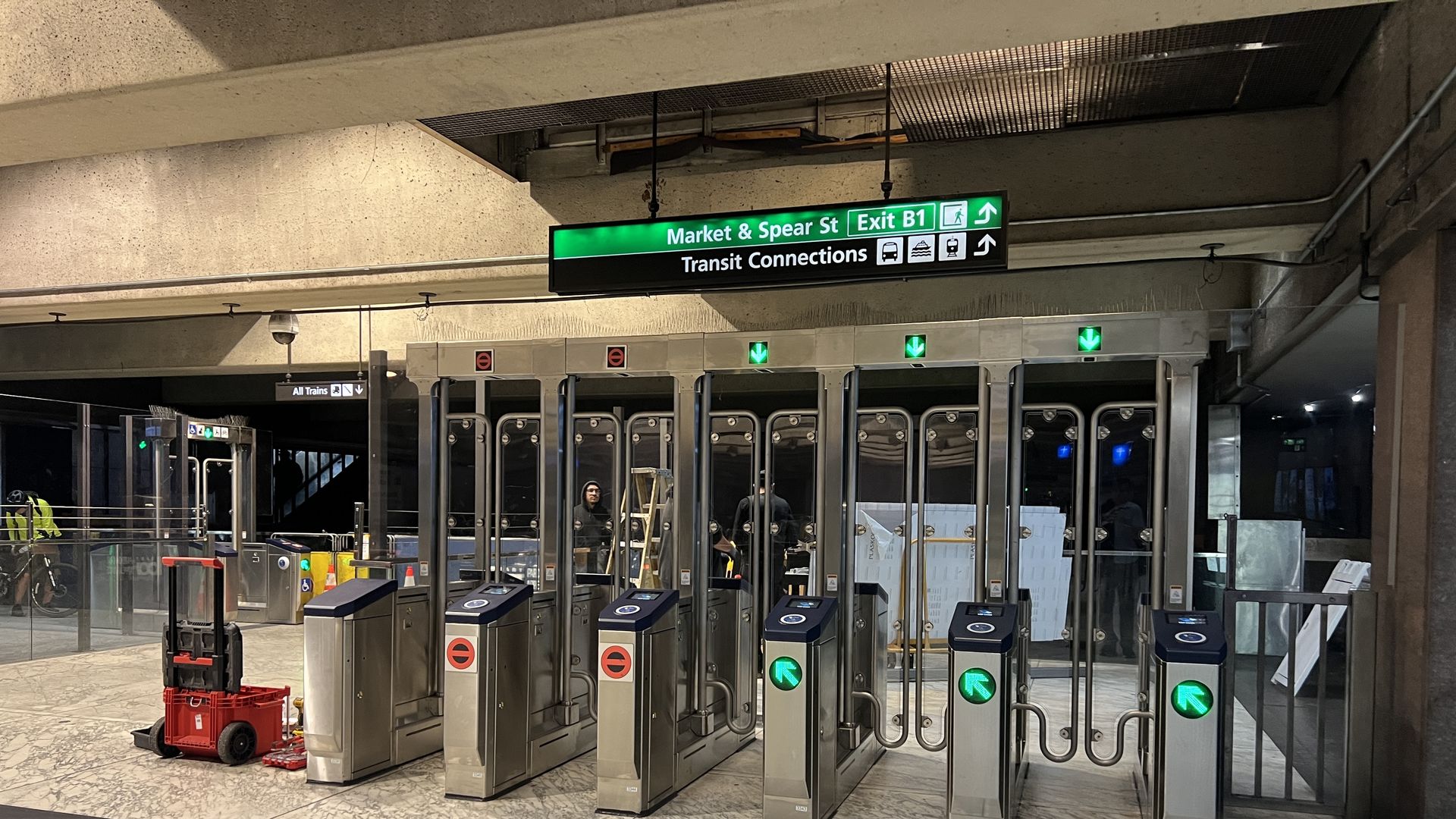 Underground transit station with metal turnstiles, green exit arrows, and a sign reading Market & Spear St Exit B1 showing transit icons. Marble floor and concrete walls; a red tool cart on the left.