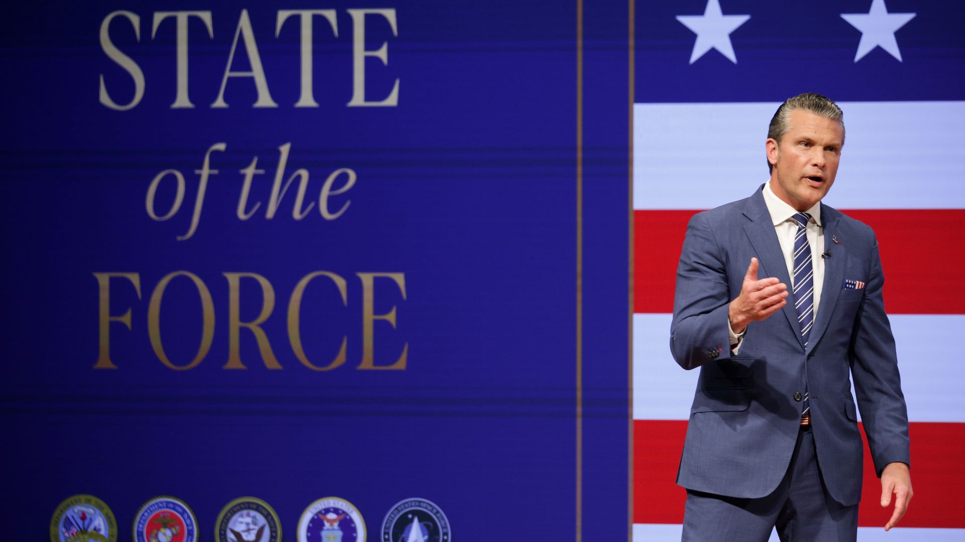 A man in a blue suit speaks on a stage with a deep blue backdrop reading "STATE of the FORCE" in gold, next to a vertical US flag panel; a row of circular emblems sits along the bottom.