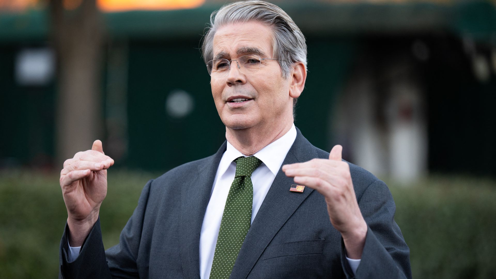 Treasury Secretary Scott Bessent, in a dark suit, white shirt and green tie, speaks to reporters while gesturing with both hands as if to describe a level. 