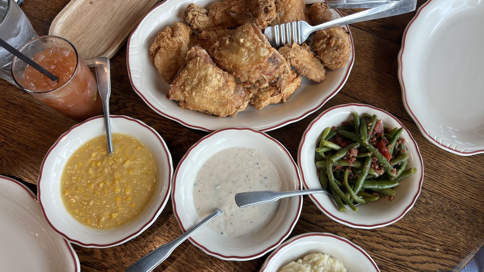Large platter of fried chicken on a red-rimmed plate, with sides of corn soup, gravy, green beans with bacon, mashed potatoes, and deviled eggs on a wooden table with drinks nearby.