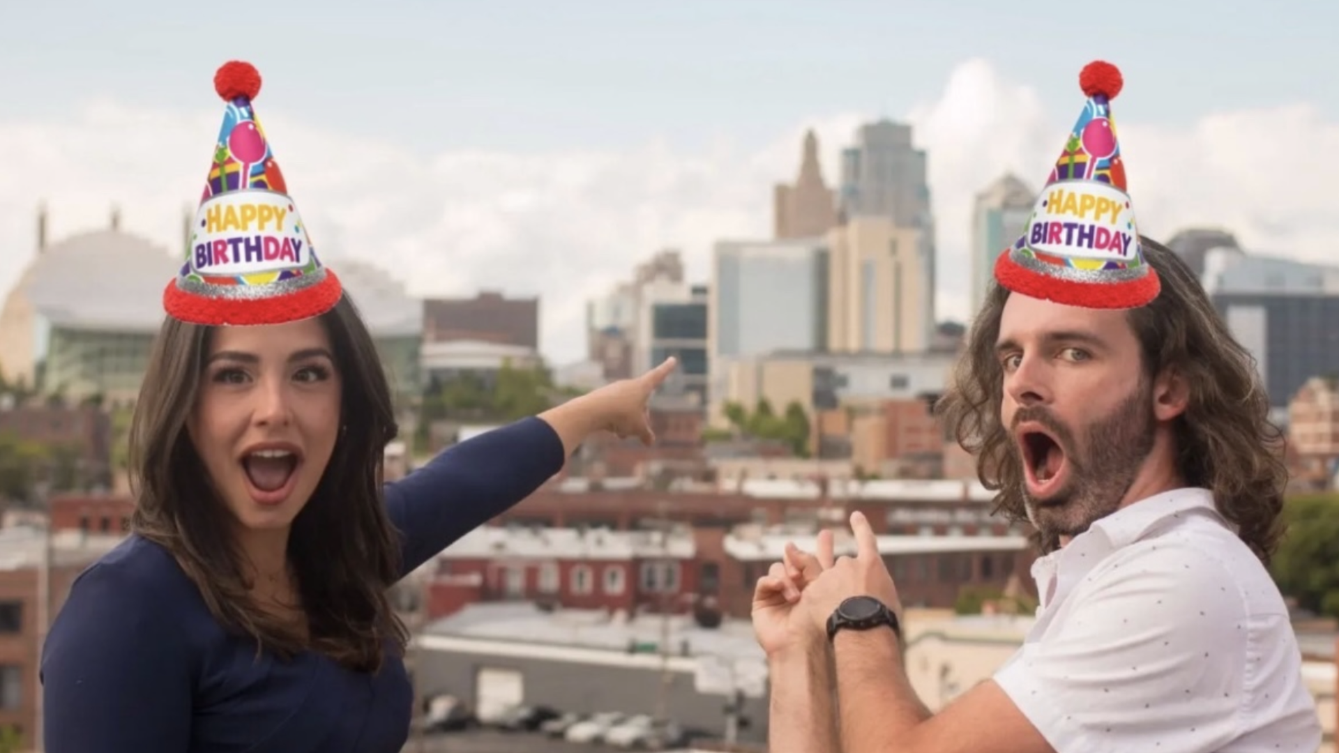Two people on a rooftop with a city skyline in the background, wearing colorful birthday hats with red pom-poms. The woman points excitedly to the left while the man looks surprised.