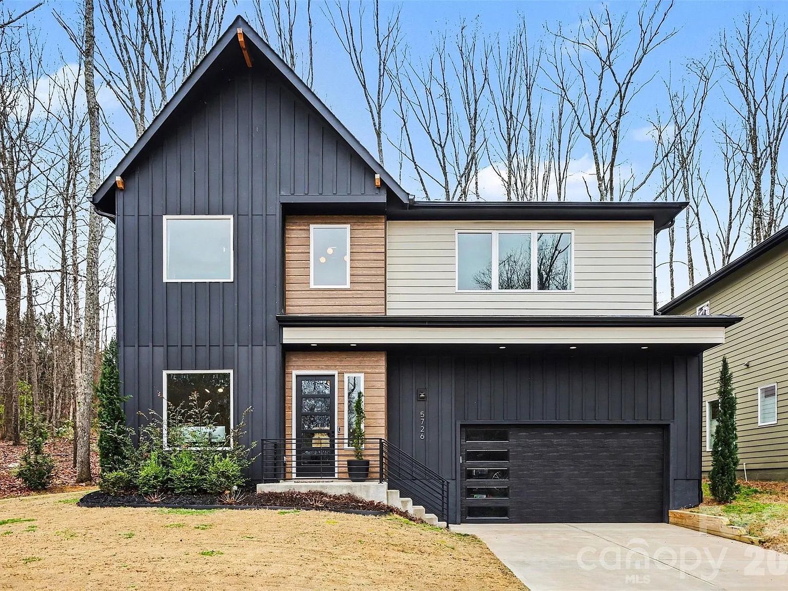 Modern two-story house with dark and light gray siding, wood accents around the front door, tall windows, black garage door, and a sloped front yard with sparse grass and trees in the background.