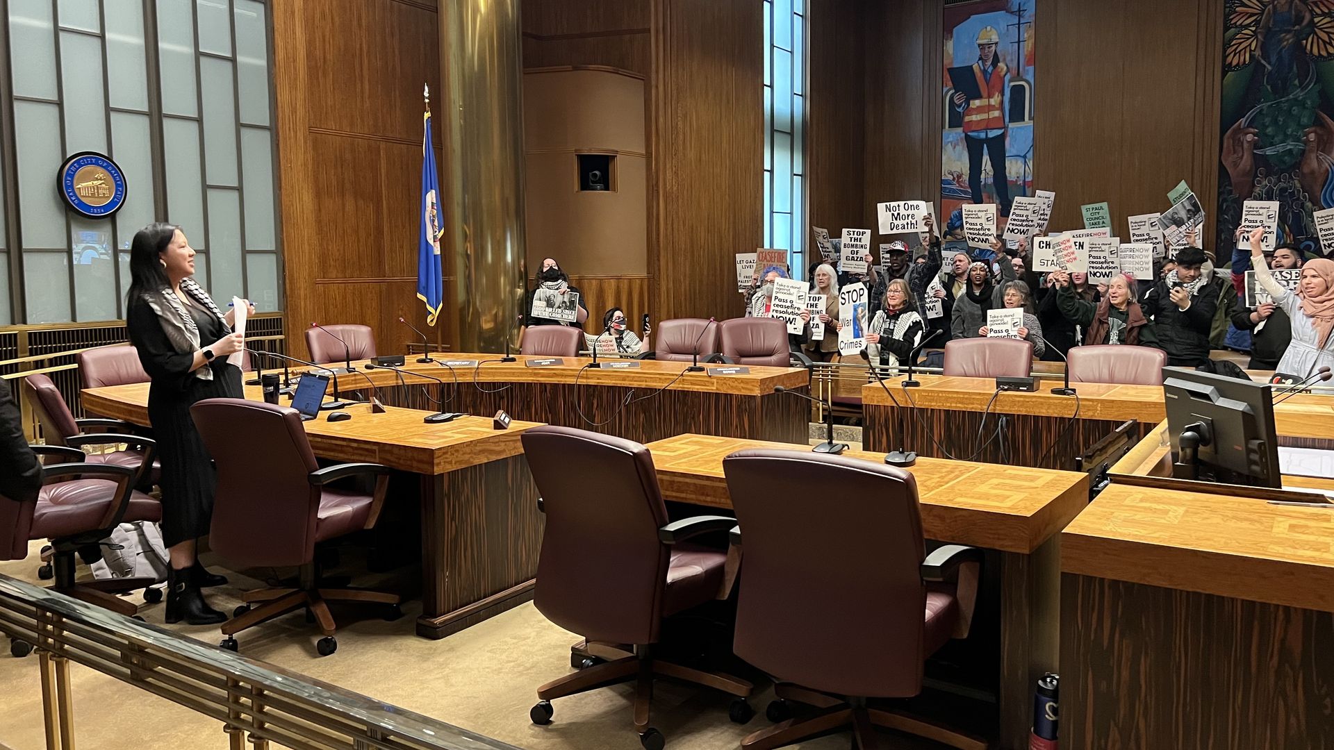 A city council chamber filled with protestors holding signs, some of which say "Ceasefire Now," with an empty dais and one woman in black clothing speaking to the crowd.