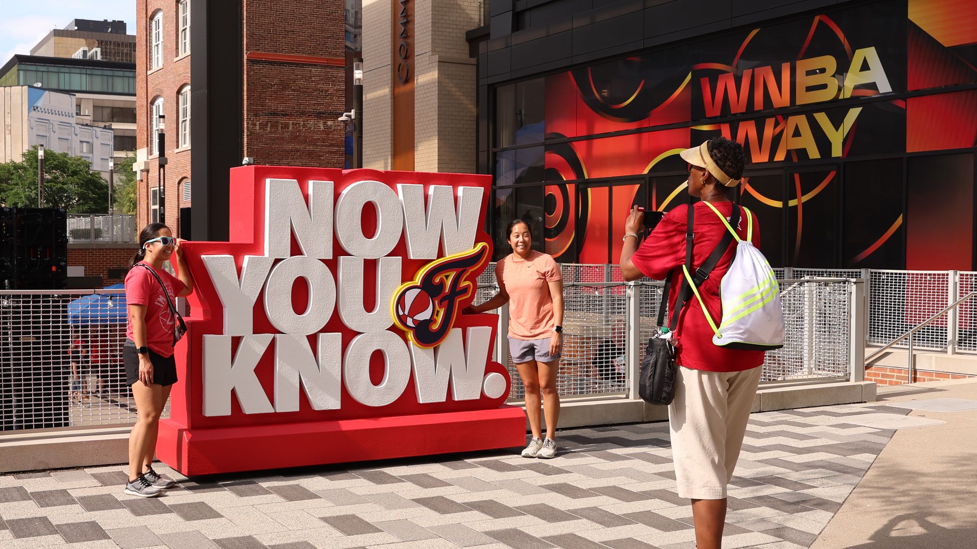 Three women outdoors near a red and white sign that says "NOW YOU KNOW" with a sports logo. One woman takes a photo of two others posing beside the sign. Bright, sunny day.