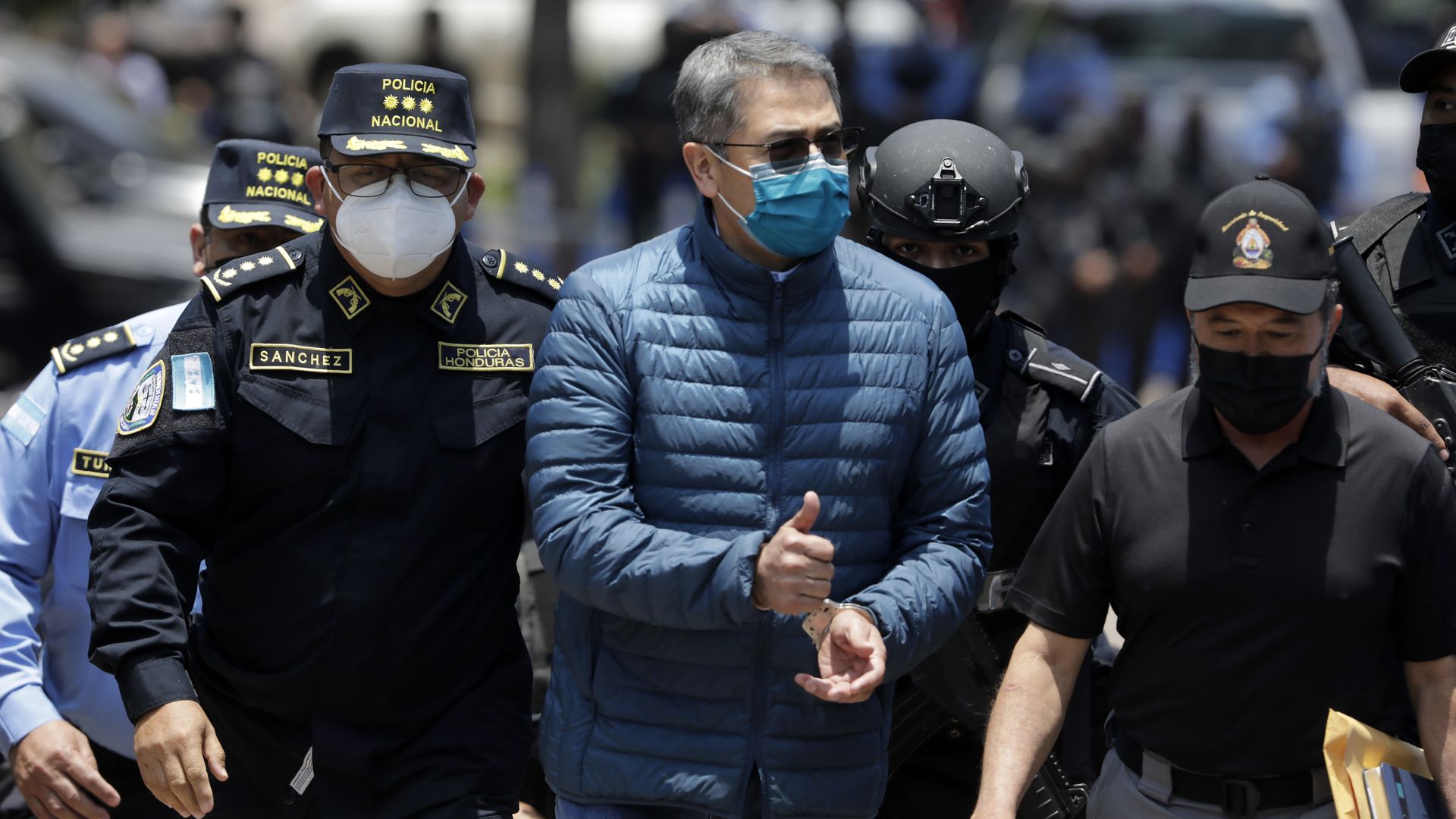 former Honduras president, wearing a blue puffer jacket, sunglasses, a blue medical mask and apparently handcuffed at the front, walks surrounded by police while being extradited