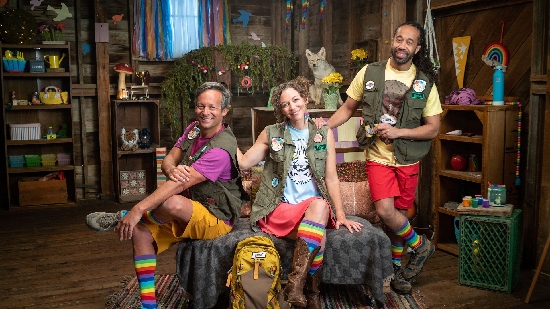 Three adults in colorful outdoor vests and rainbow socks pose smiling inside a rustic room decorated with rainbows, plants, and crafts. A yellow backpack sits in front.