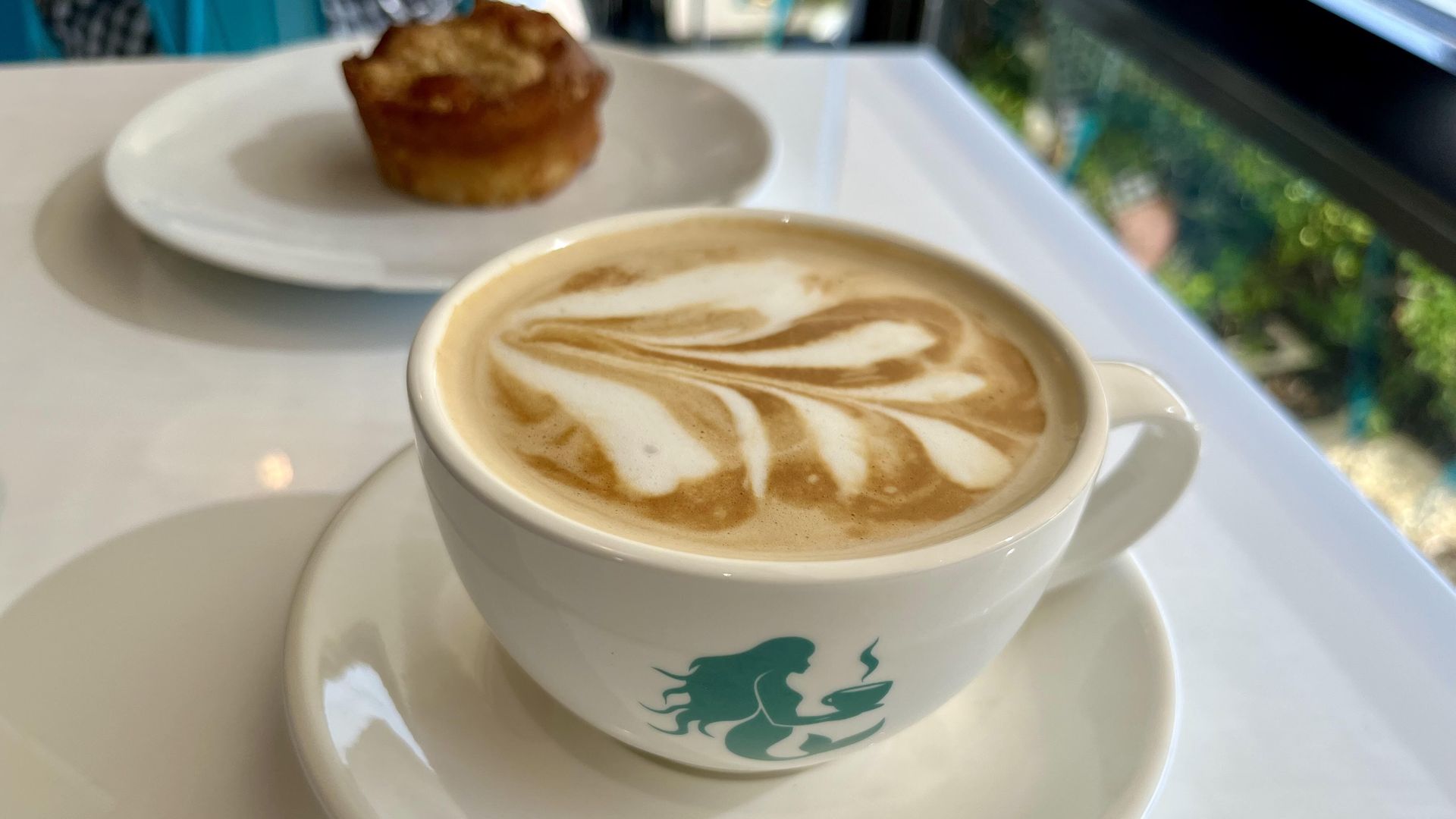Close-up of a white cup with latte art, featuring a brown and white leaf pattern on top. The cup has a green mermaid logo, placed on a white saucer on a white table with a blurred pastry in the background.