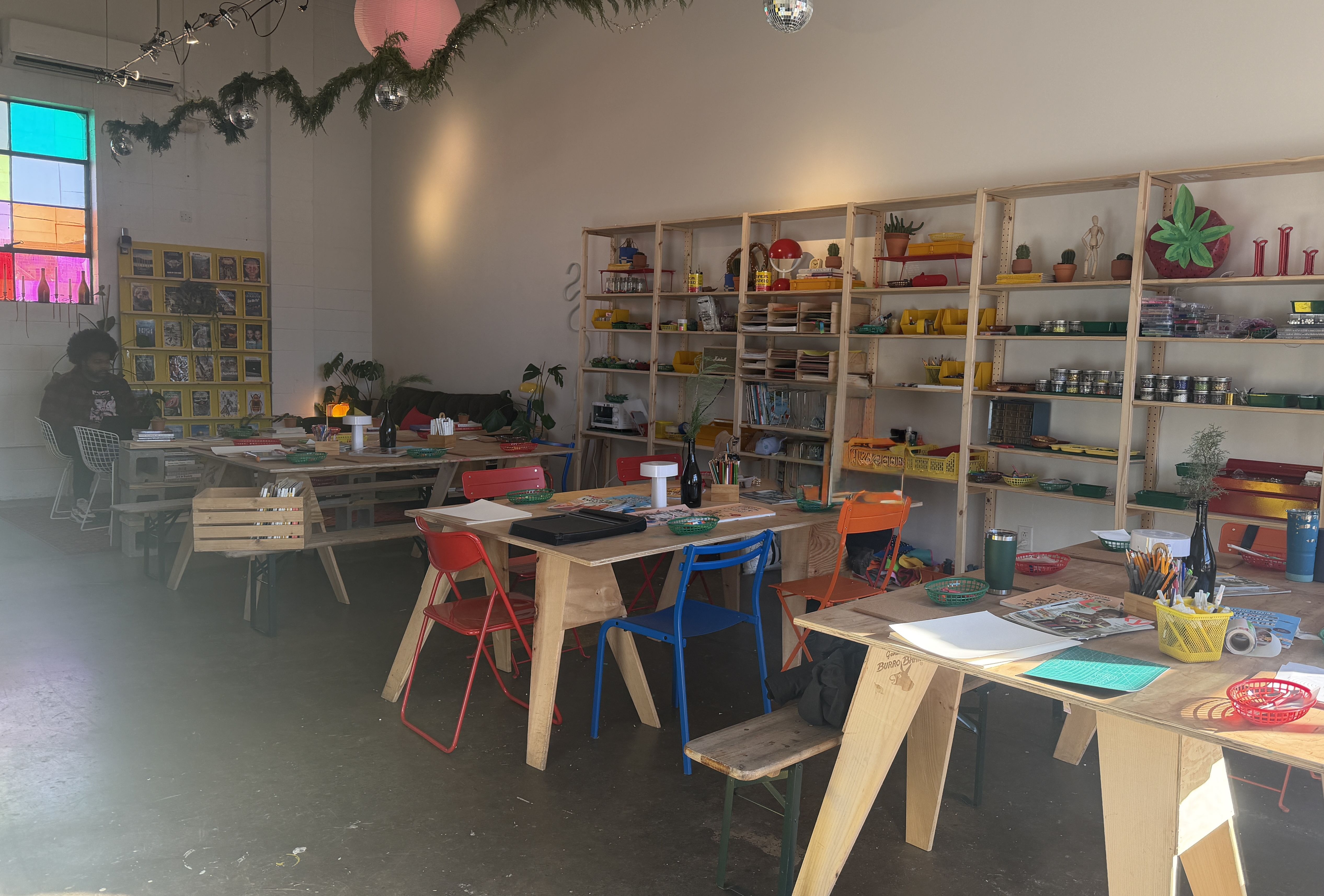Bright, colorful art studio with wooden tables, blue, red, orange chairs, craft supplies, plants, open shelving, a stained glass window, and a person sitting in the back corner.