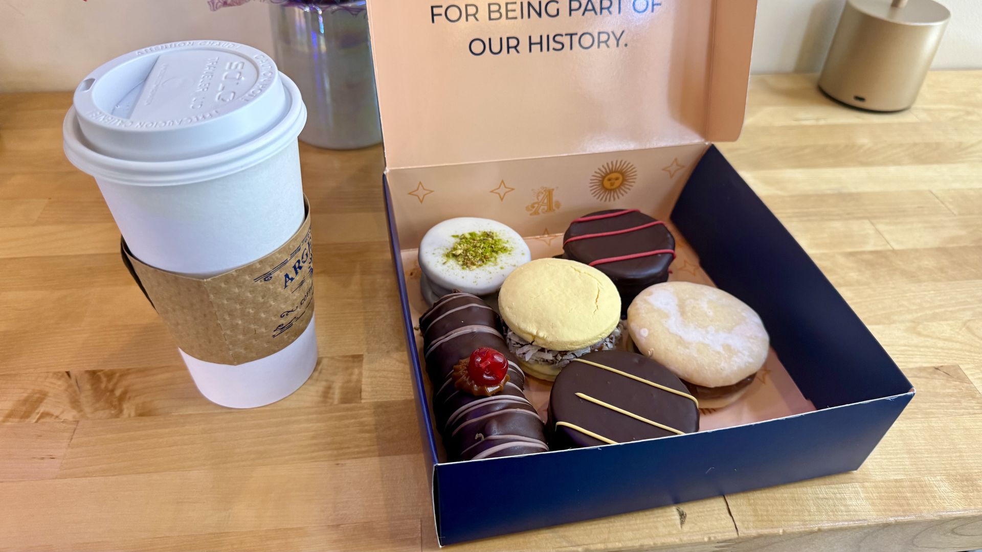 Open box of assorted donuts on a wooden table beside a white takeaway coffee cup. Frosted and chocolate-dipped donuts, including pistachio-topped varieties, are visible inside.