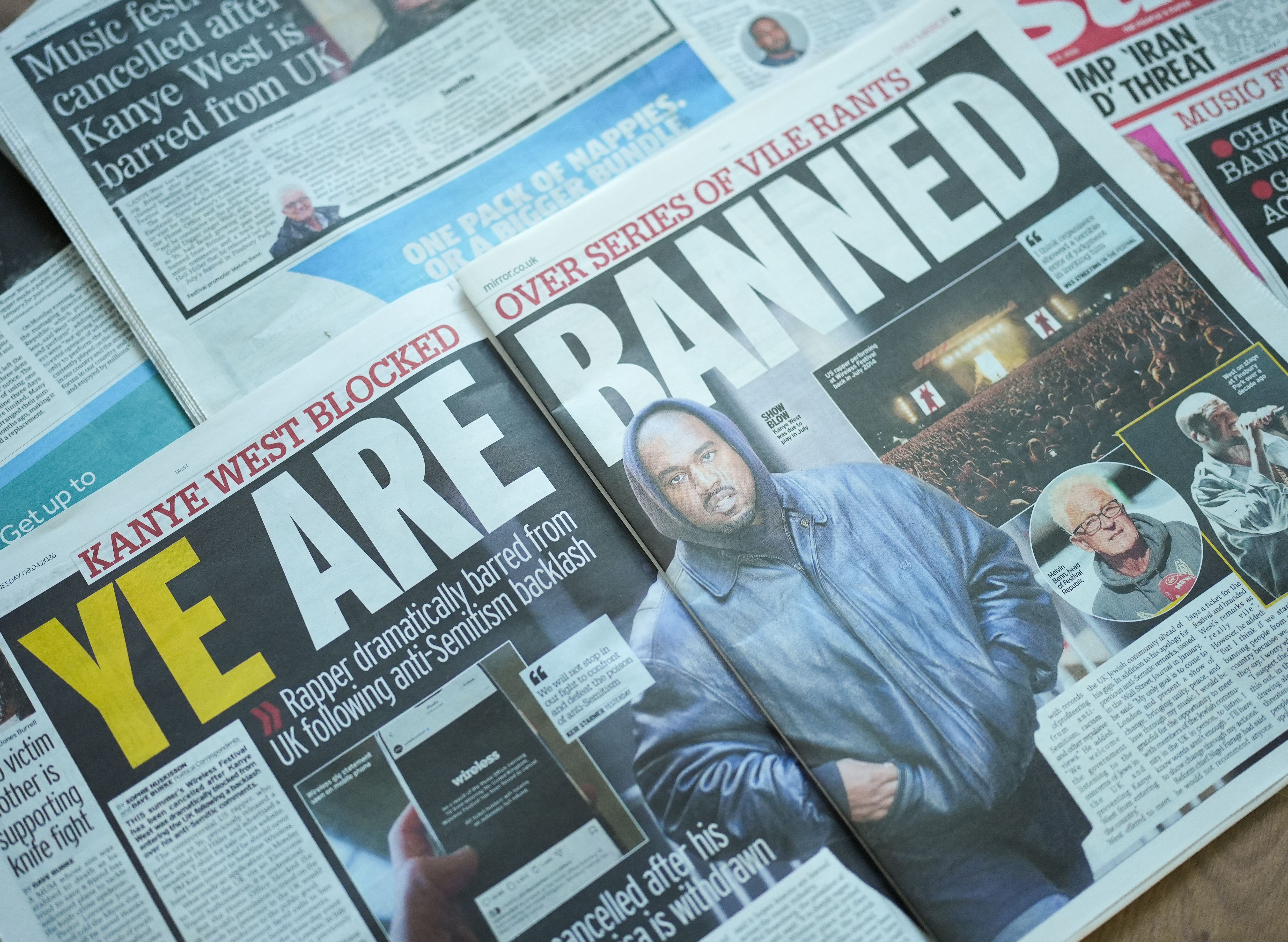 A photo of open tabloids spread on a flat surface. The center one shows a picture of Ye with the headline "Ye Are Banned."