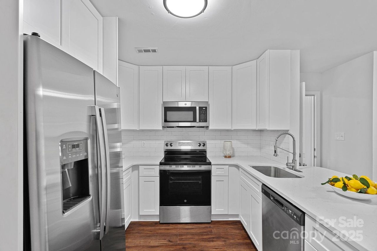 Modern white kitchen with stainless steel refrigerator, oven, microwave, and dishwasher. White countertops, subway tile backsplash, wood floor, and a plate of yellow lemons on the counter.