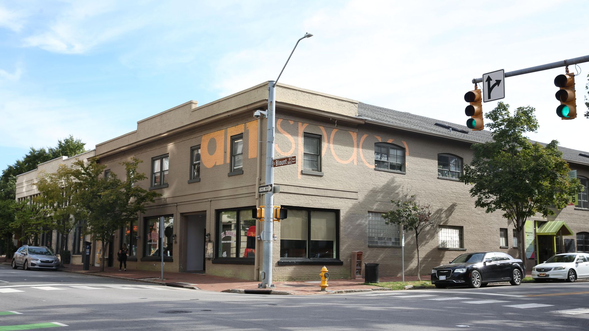 Corner building with beige brick exterior and orange partial text "art space," trees lining the sidewalk, parked cars, traffic lights, and street signs on a sunny day.
