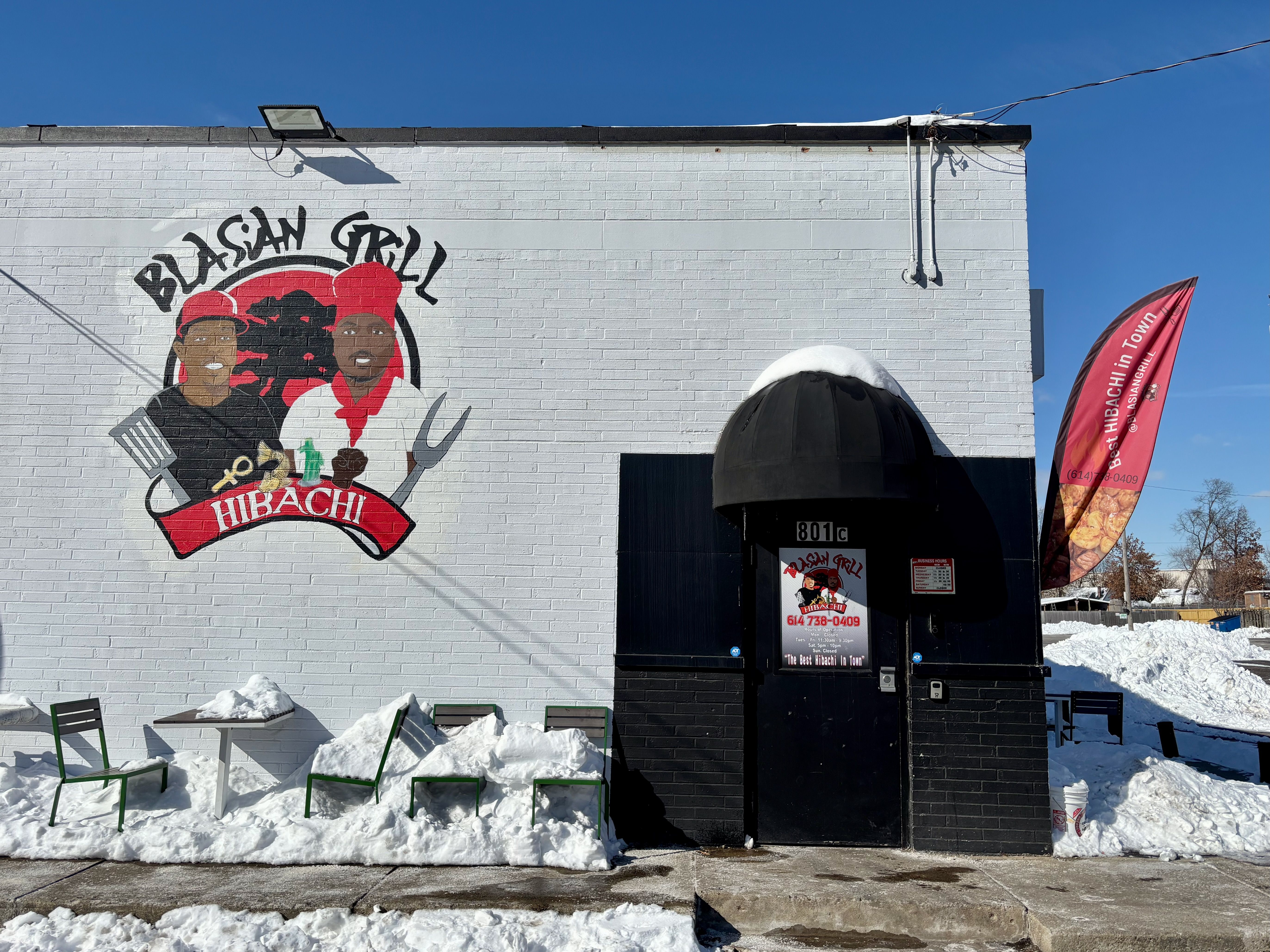 Exterior of Blasian Grill Hibachi with a mural of two chefs holding grill tools on a white brick wall. Snow covers outdoor tables and chairs, clear blue sky above.
