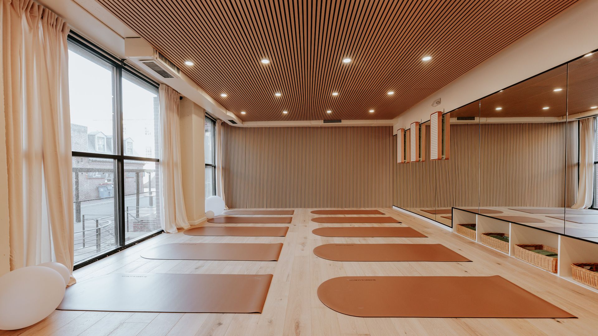 Bright yoga studio with light wood floor and ceiling, brown mats neatly arranged, large windows with cream curtains, and a mirrored wall reflecting the space.