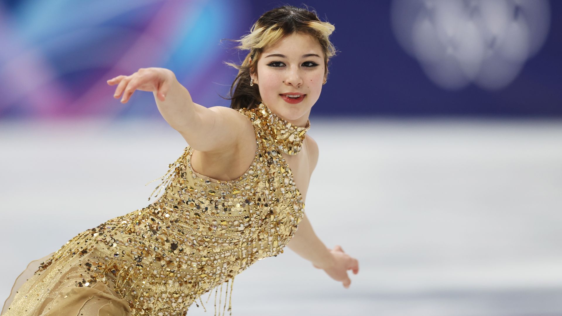 A female figure skater in a gold sequined dress extends her arm while skating on ice; a blurred blue background with soft bokeh lights.