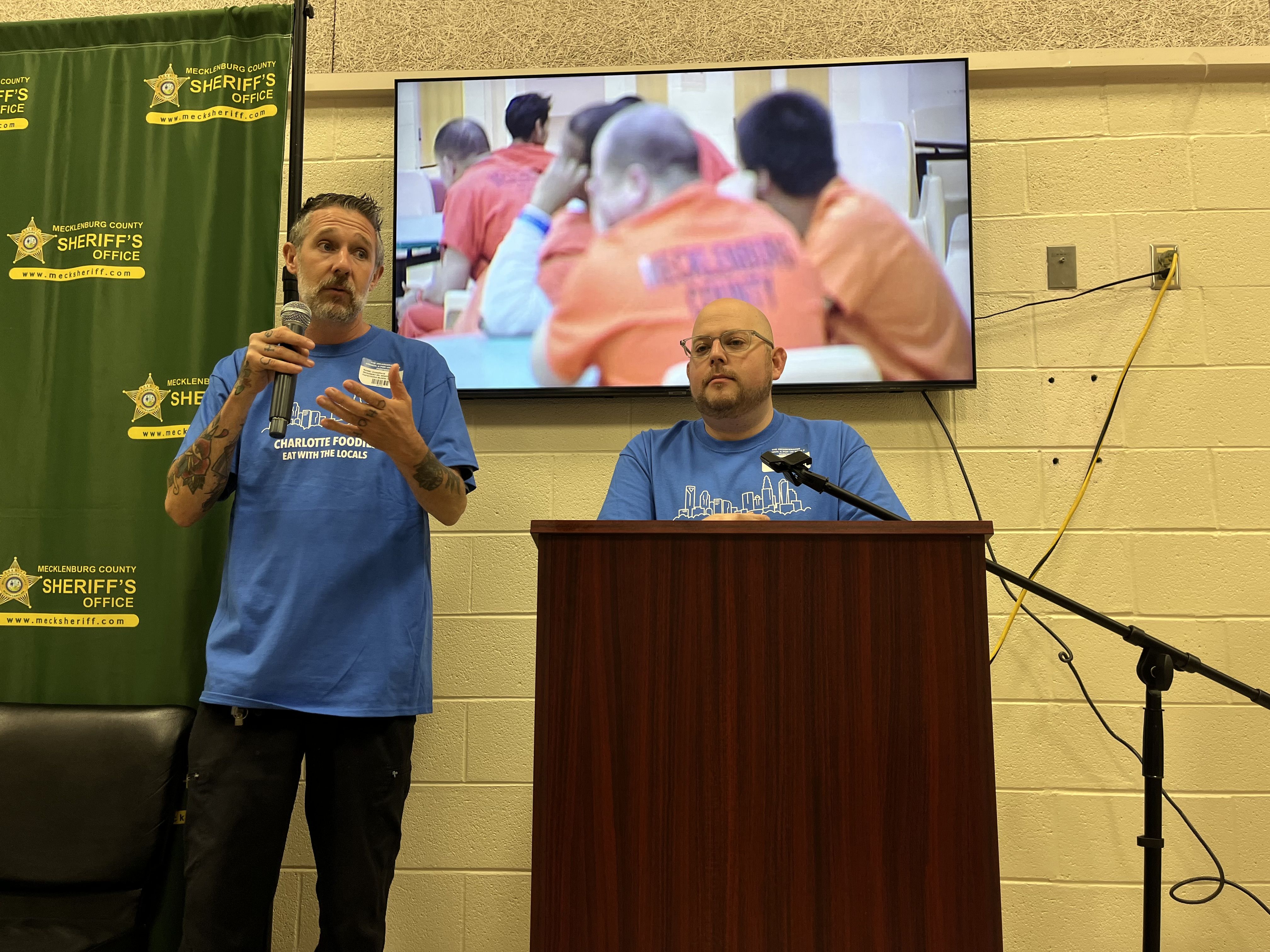 Two men wearing blue "Charlotte Foodie" T-shirts speak at a podium with a microphone; behind them is a screen showing people in orange prison uniforms and a green Mecklenburg County Sheriff's Office banner.