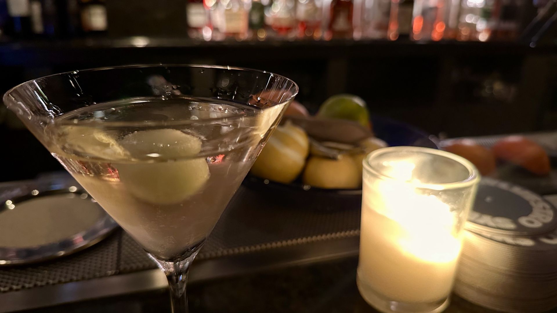 A close-up of a clear martini glass with two olives inside, lit softly by a glowing candle on the right. Blurry background shows bottles and citrus fruits on a bar counter.