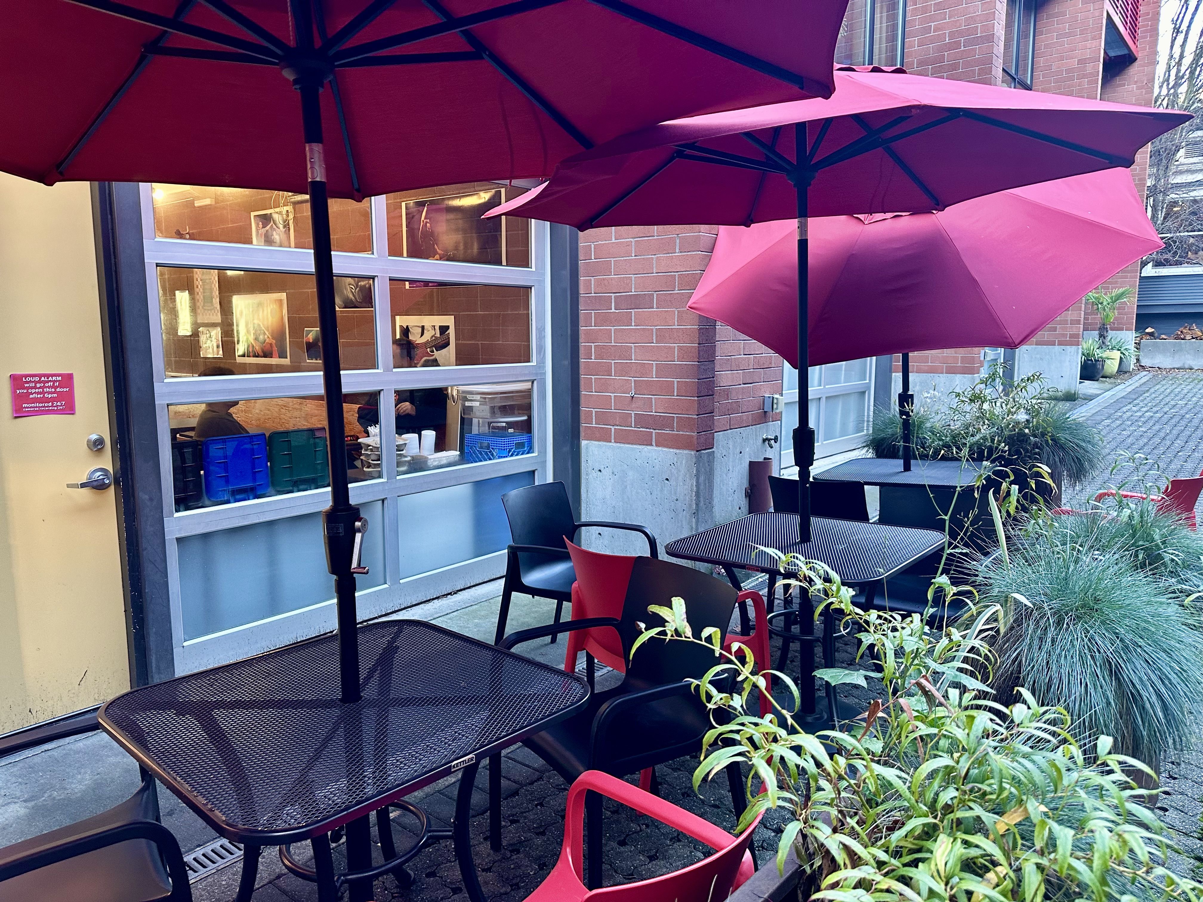 Red umbrellas above outdoor tables, in front of a garage-style window that leads to an indoor cafe.