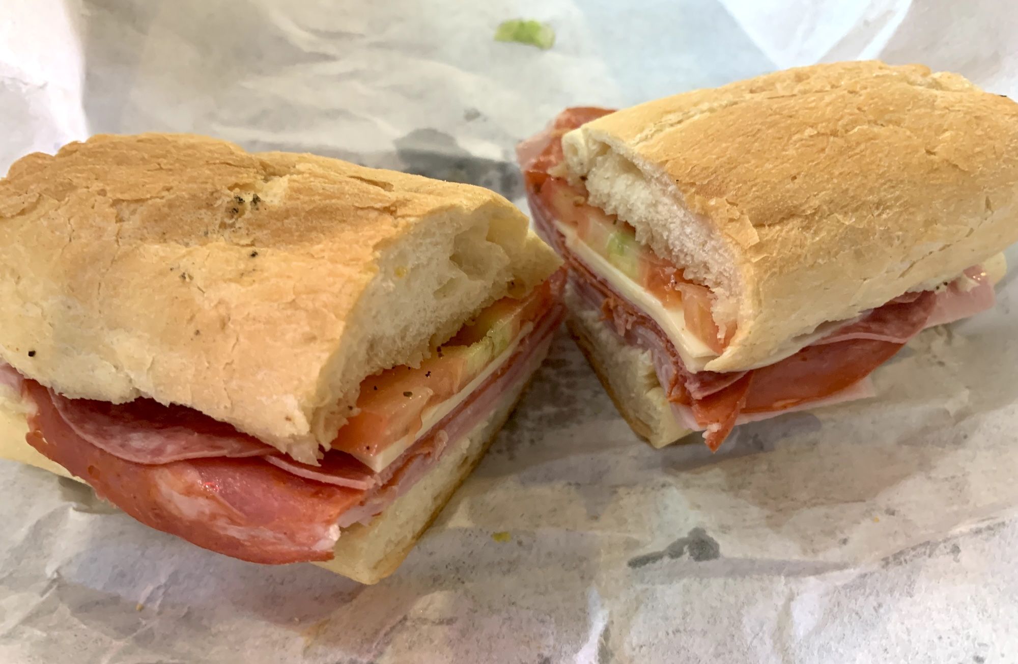 Two halves of a crusty baguette sandwich filled with ham, cheese, tomato, and lettuce on pale white bread, resting on parchment paper.