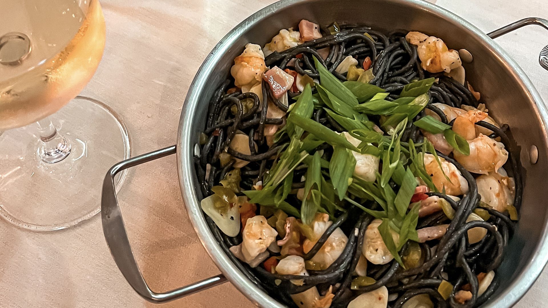 A table setting with a silver bowl of black pasta and a glass of white wine.